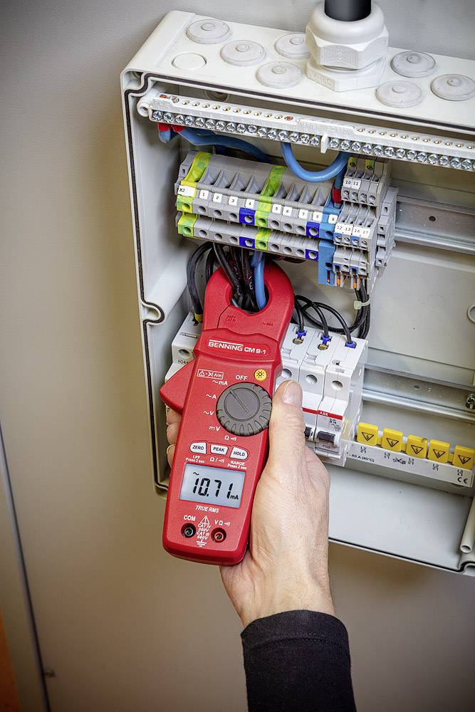 Osoba používá červený ampérmetr ke kontrole elektrických kabelů v otevřené rozvodné skříni.