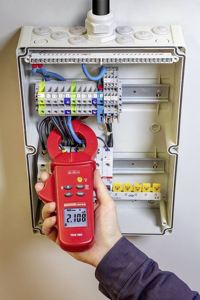 Jedna osoba měří elektrický proud v otevřené rozvodné skříni pomocí červených kleští. Displej ukazuje '2,108'.