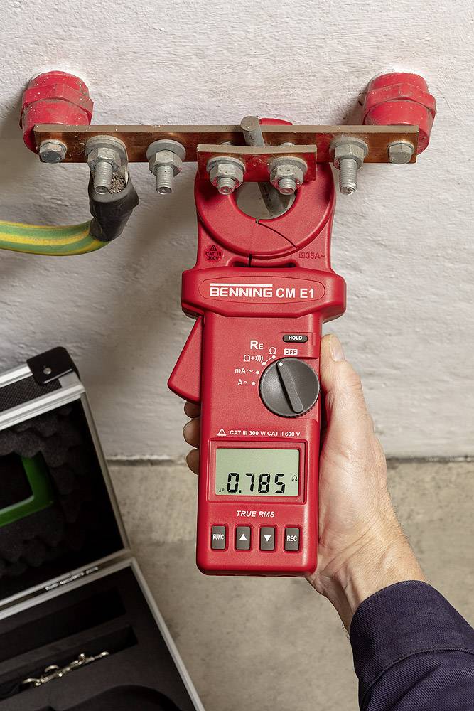 Osoba měří elektrický proud na vedení červeným digitálním měřicím přístrojem. Display ukazuje '0.785'.