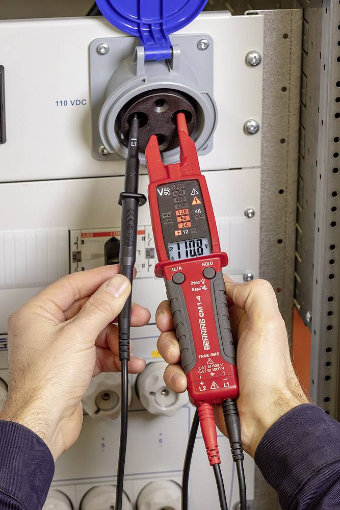 Člověk měří napětí v elektrické zásuvce pomocí digitálního voltmetru. Na displeji je zobrazeno 110 V DC.