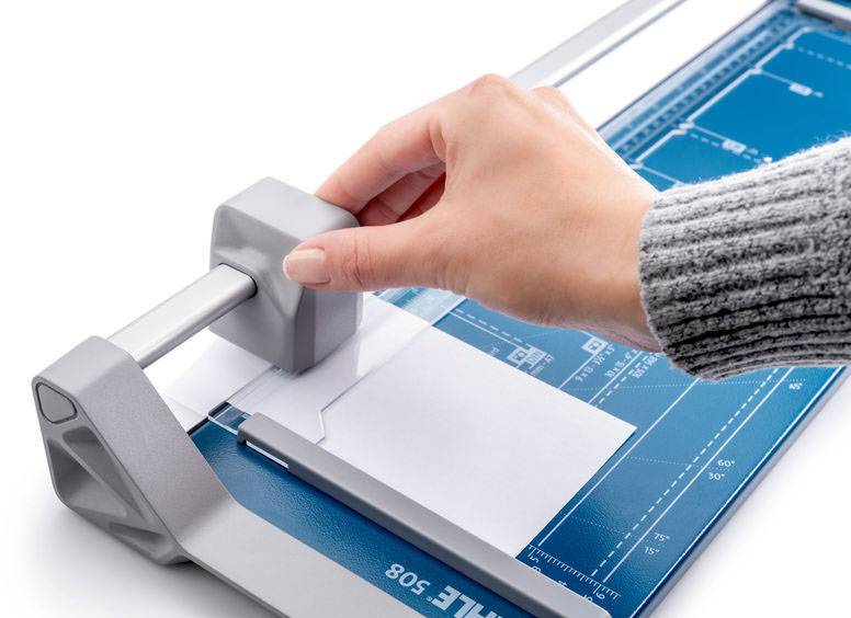 Ruka řeže list papíru pomocí řezačky. Řezací stůl má vyznačené linky pro přesné zarovnání.