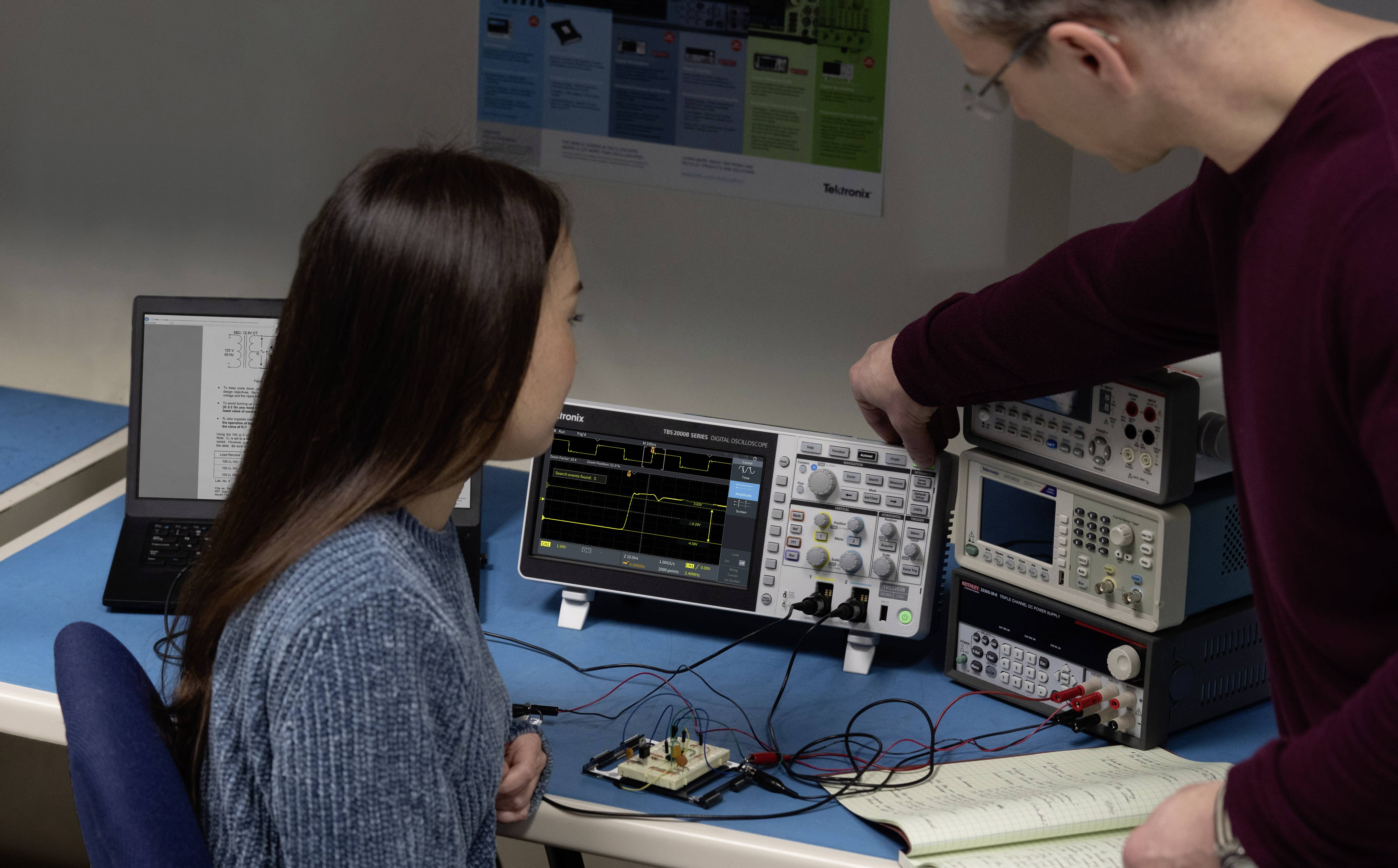 Muž vysvětluje ženě použití osciloskopru. V pozadí jsou viditelné počítače a elektronická zařízení.