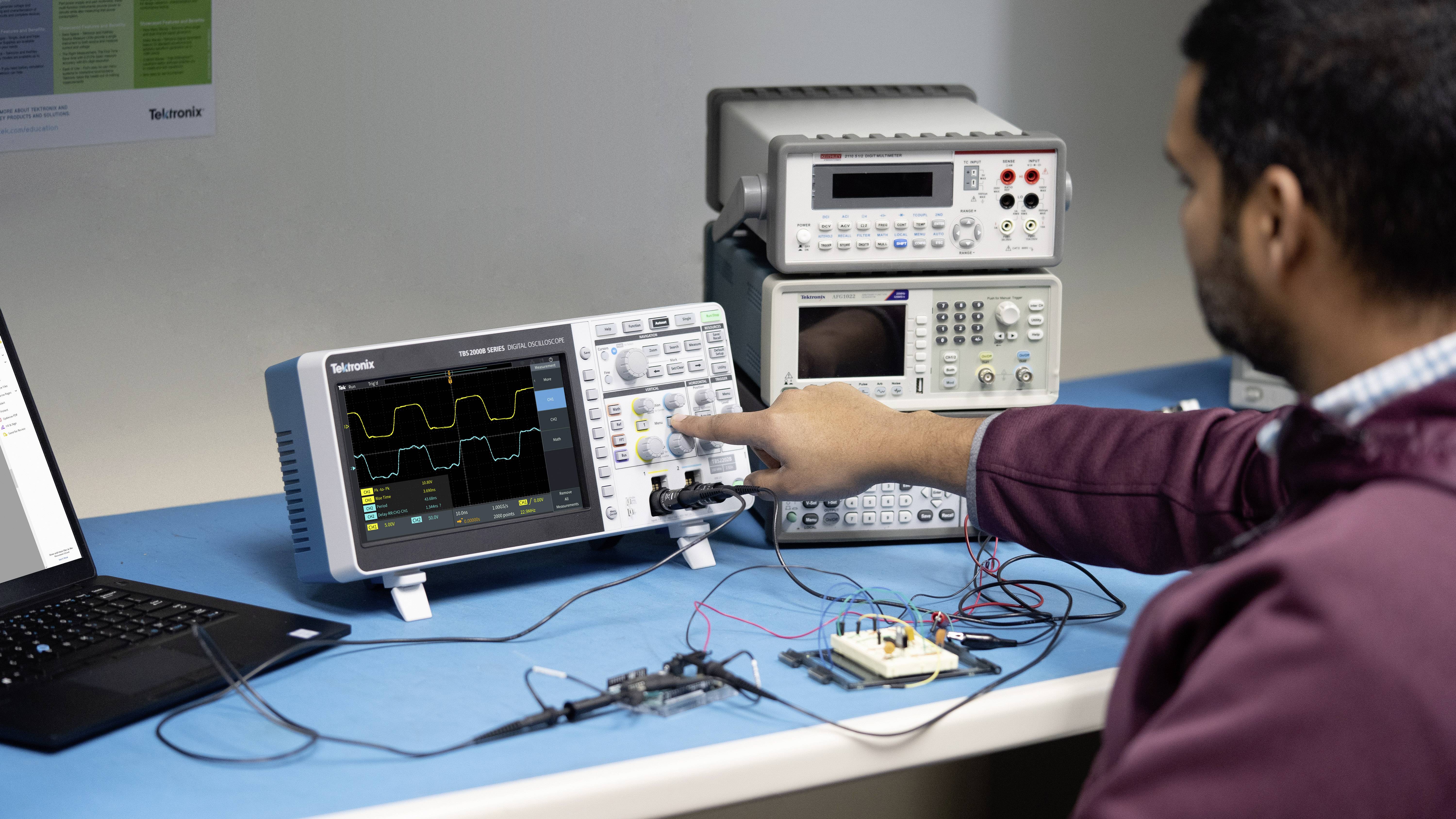 Muž obsluhuje osciloskop u stolu, který je připojený k elektronickým zařízením a laptopem.