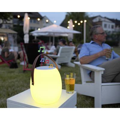 Muž sedí v zahradě a drží nápoj. V popředí svítí lampa s notami. V pozadí je uvolněná letní atmosféra.