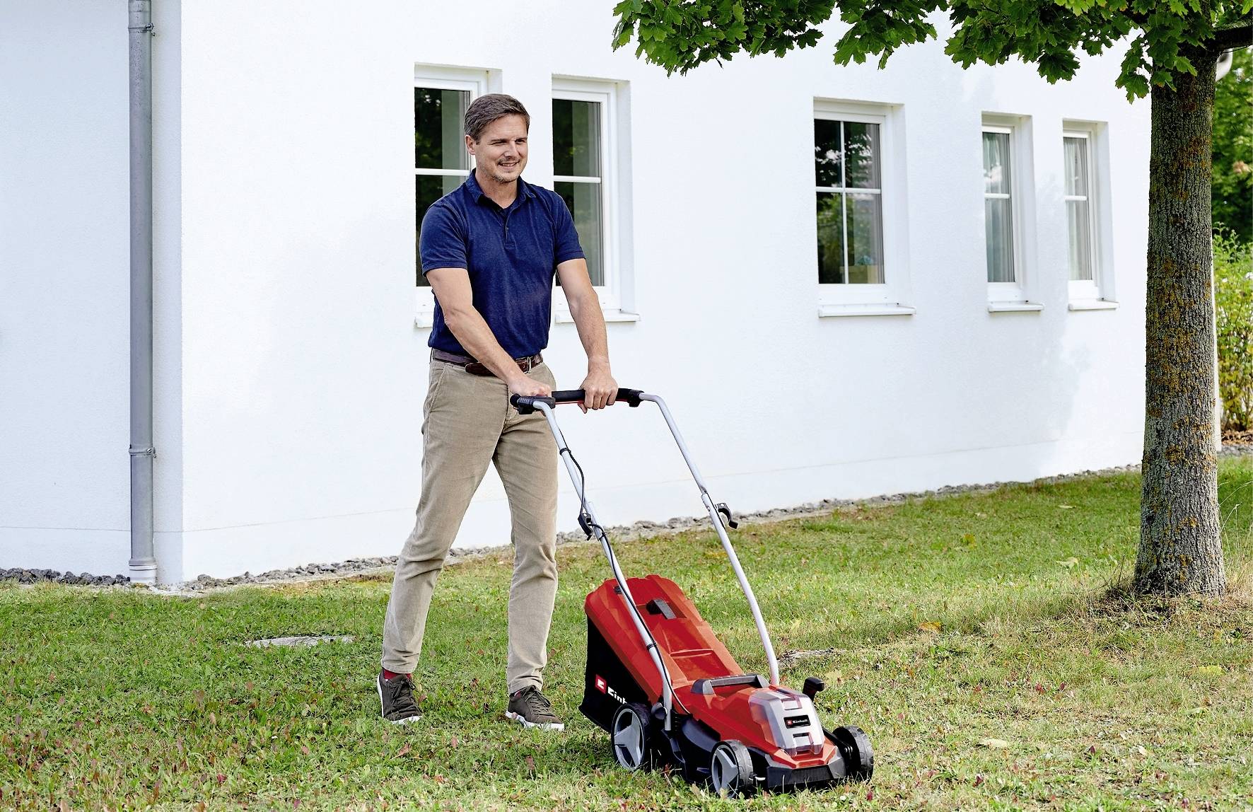 Muž seká trávu před bílým domem elektrickým, červeným sekacím strojem. Obloha je modrá, vpravo stojí strom.