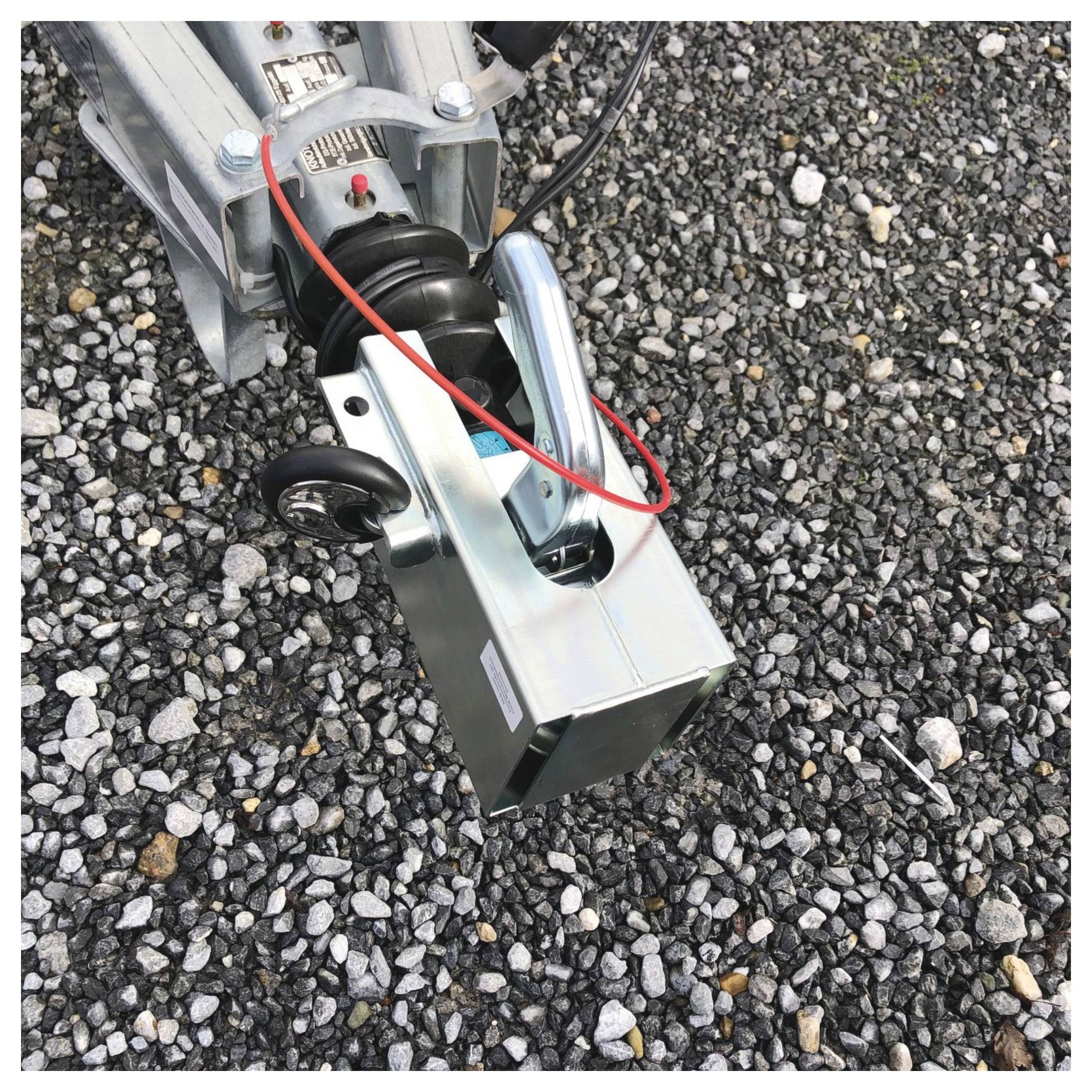 Kovový přívěsný zvedák s červenými a černými dráty odpočívá na štěrkovém povrchu. Zvedák je součástí většího mechanického zařízení.
