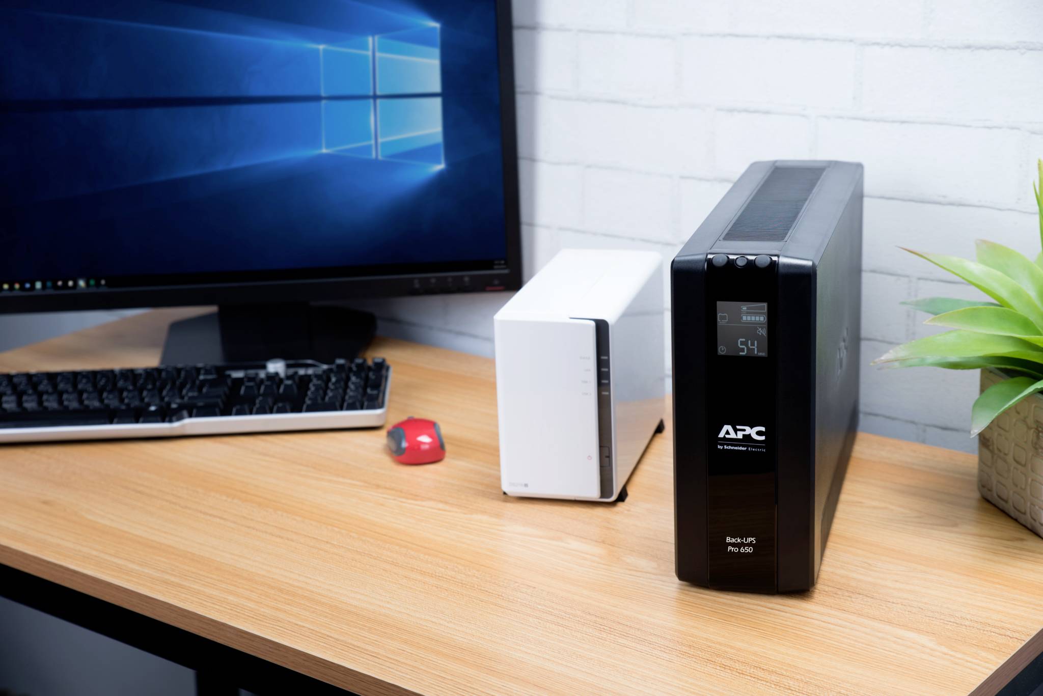 Monitor s Windows desktopem vedle záložního zdroje napájení (UPS) a bílého síťového úložiště (NAS) na pracovním stole.