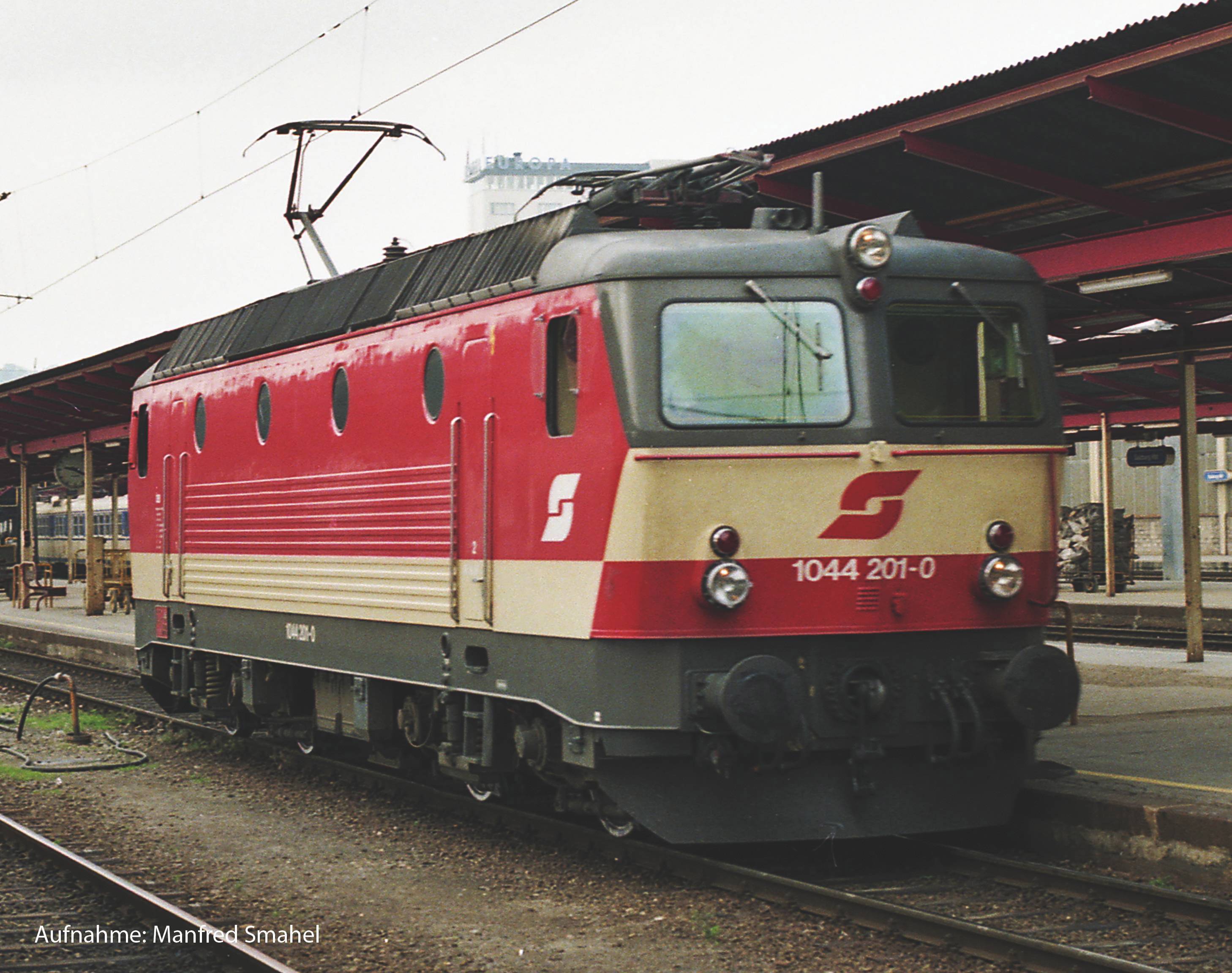 Červená a béžová elektrická lokomotiva série 1044 stojí na koleji nádražního kolejiště, obklopena krytými nástupišti.