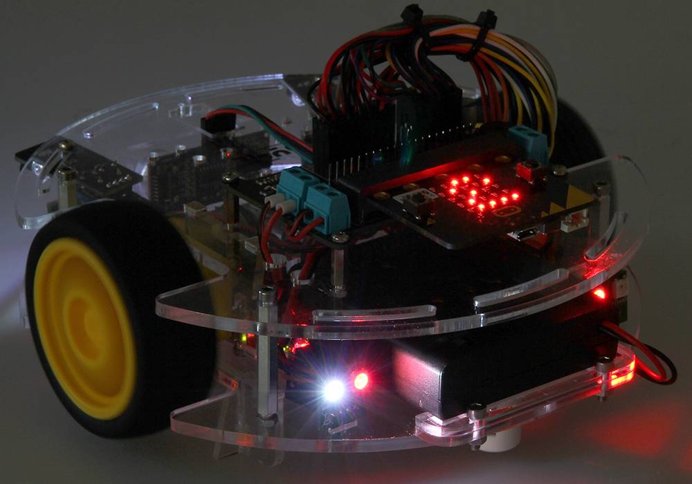 Malý programovatelný robot s žlutými koly a svítícími LED indikátory na průhledné platformě.