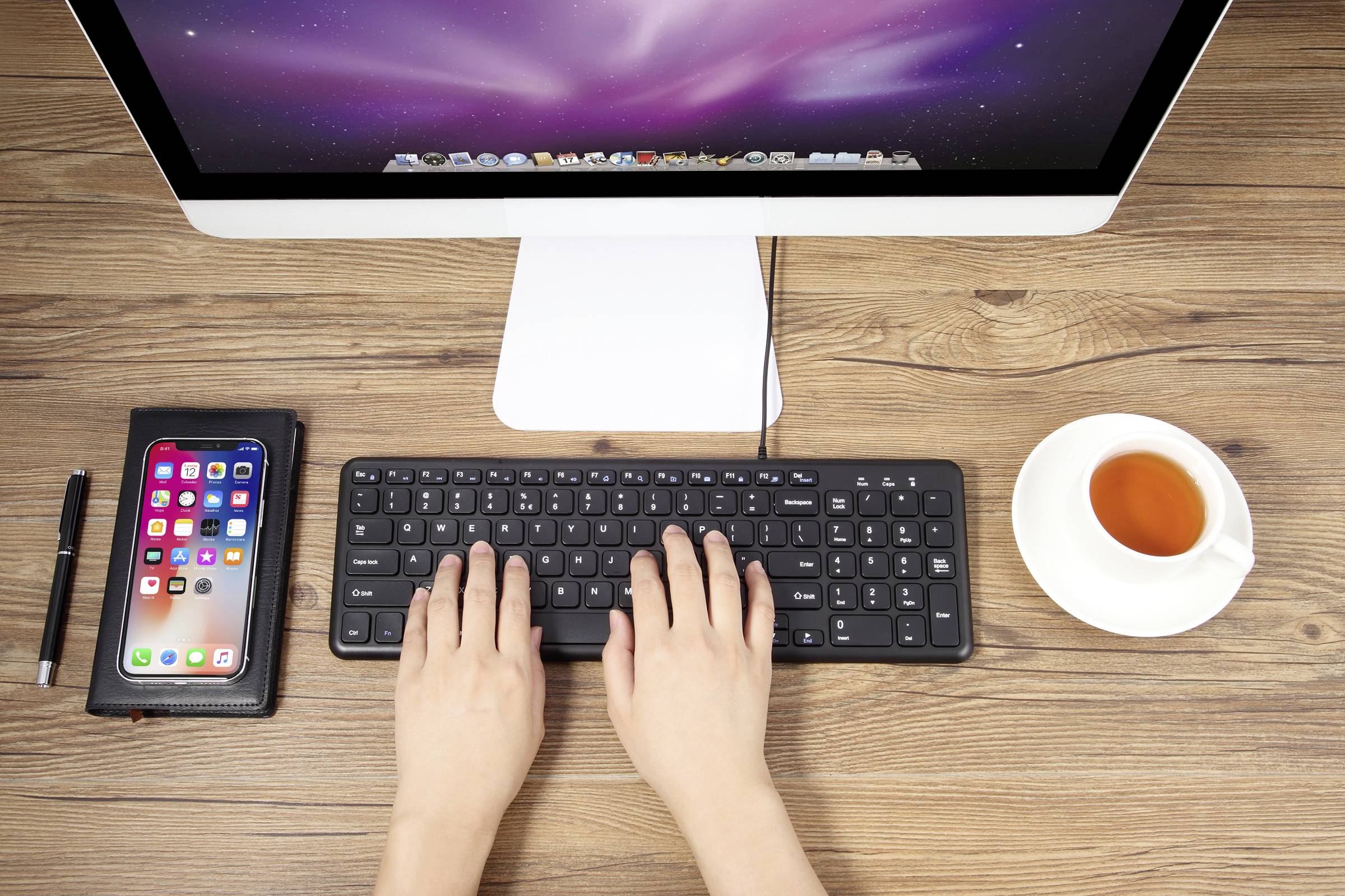 Ruce se pohybují po klávesnici před monitorem. Vedle leží smartphone, pero a stojí šálek čaje na pracovním stole.