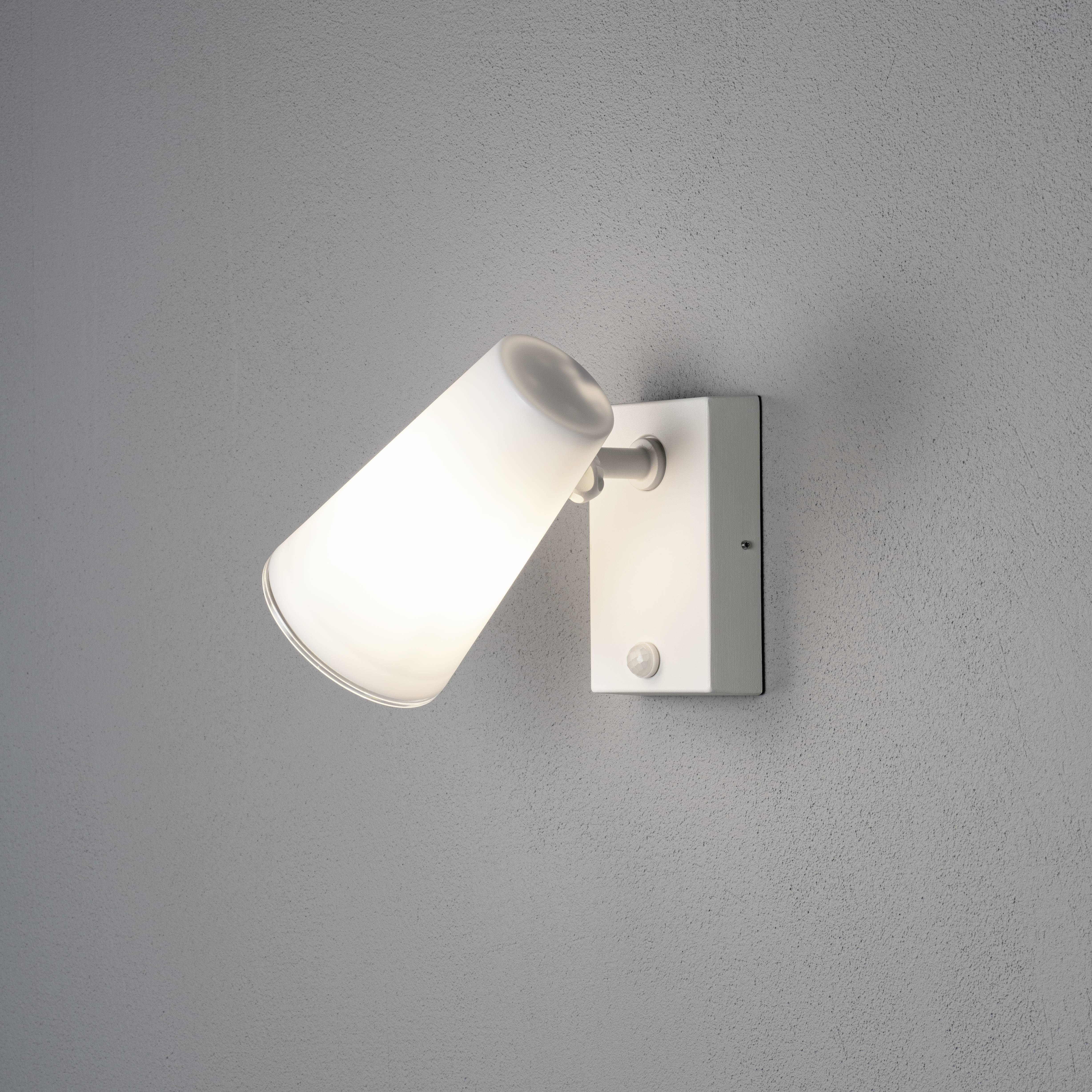 Nástěnná lampa s bílým kuželovitým stínidlem na šedé stěně, zapnutá a svítící.
