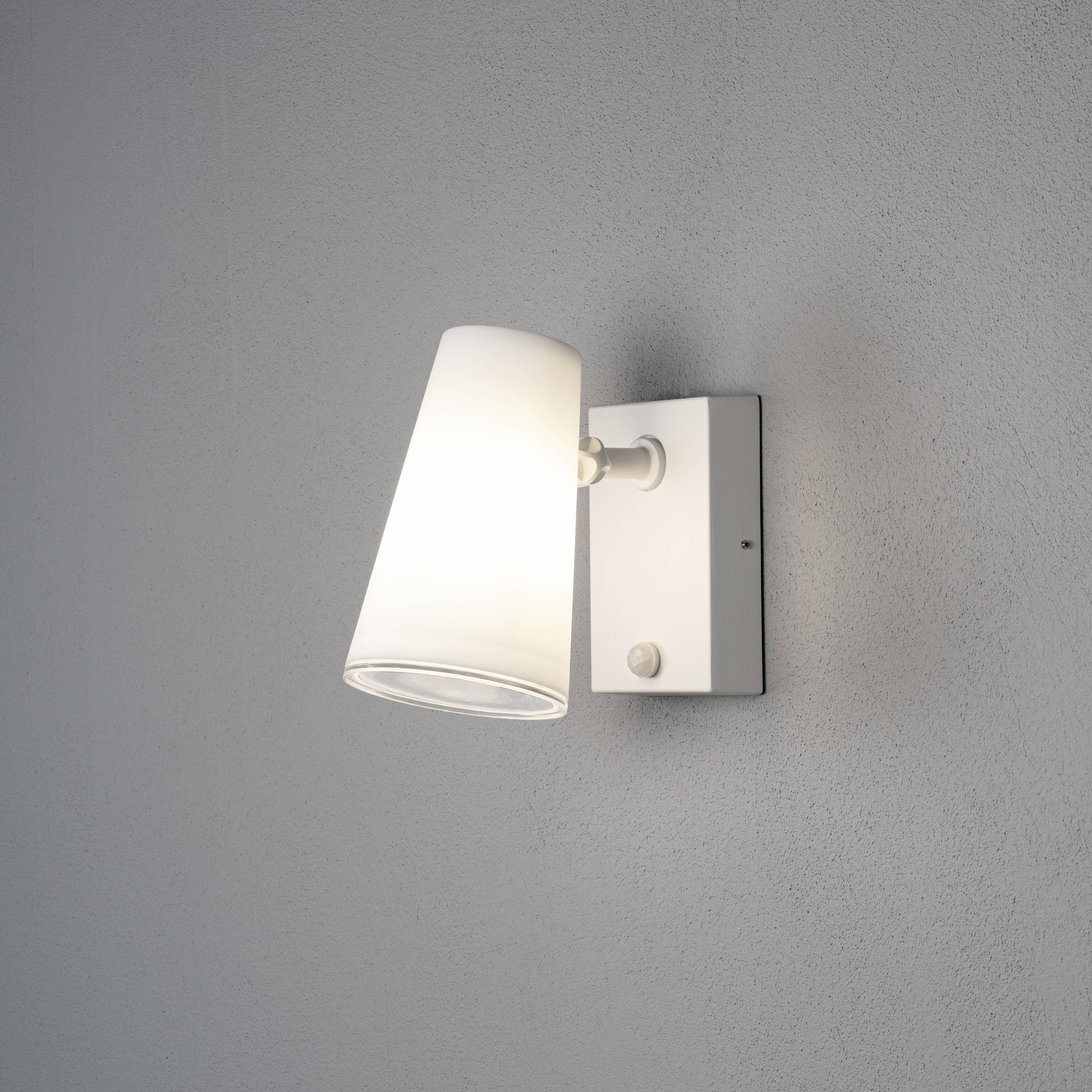 Nástěnná lampa s bílým stínidlem osvětluje měkké světlo na šedé stěně. Jednoduché, moderní designové řešení.