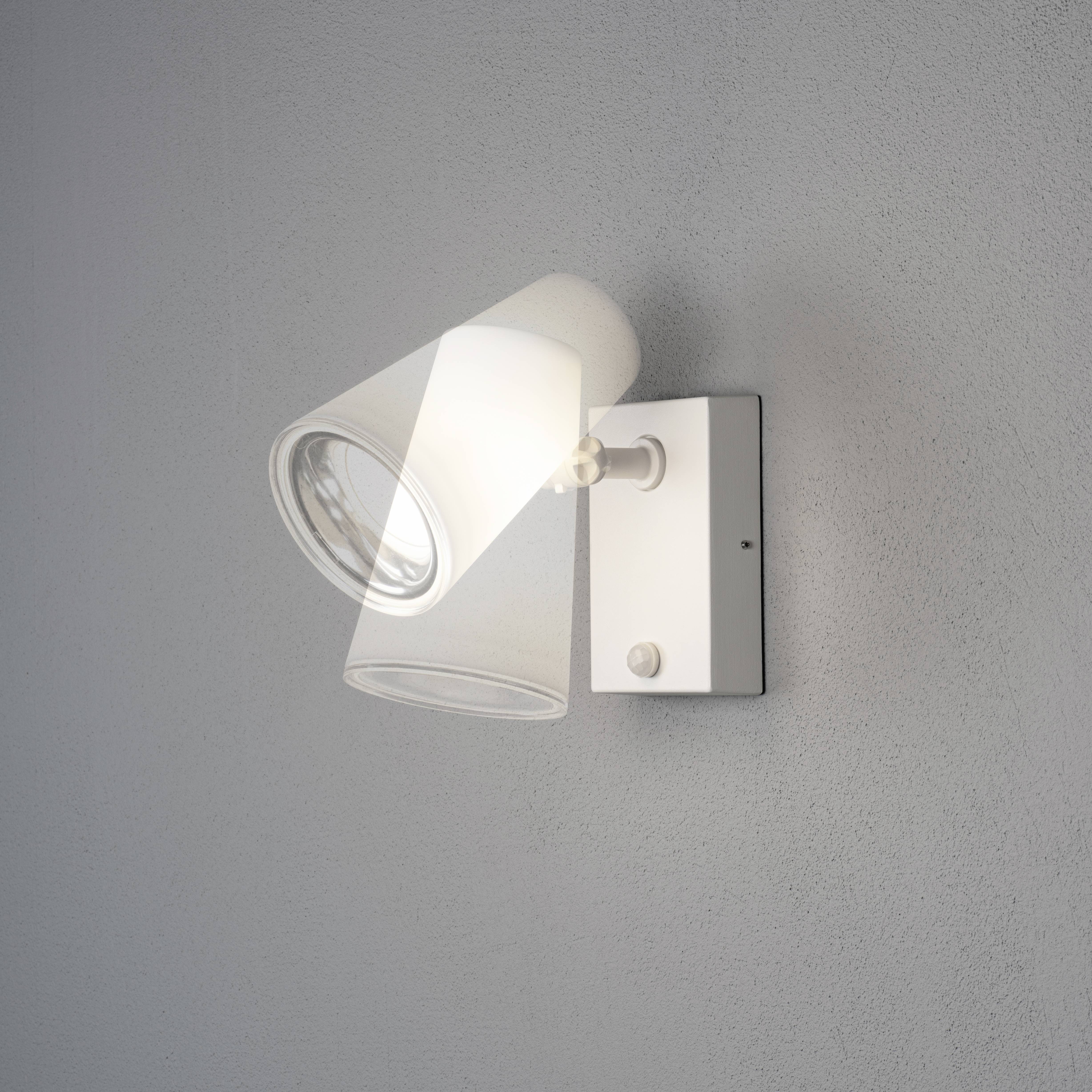 Bílá nástěnná lampa s otočným válcovitým stínidlem, namontovaná na šedé zdi.