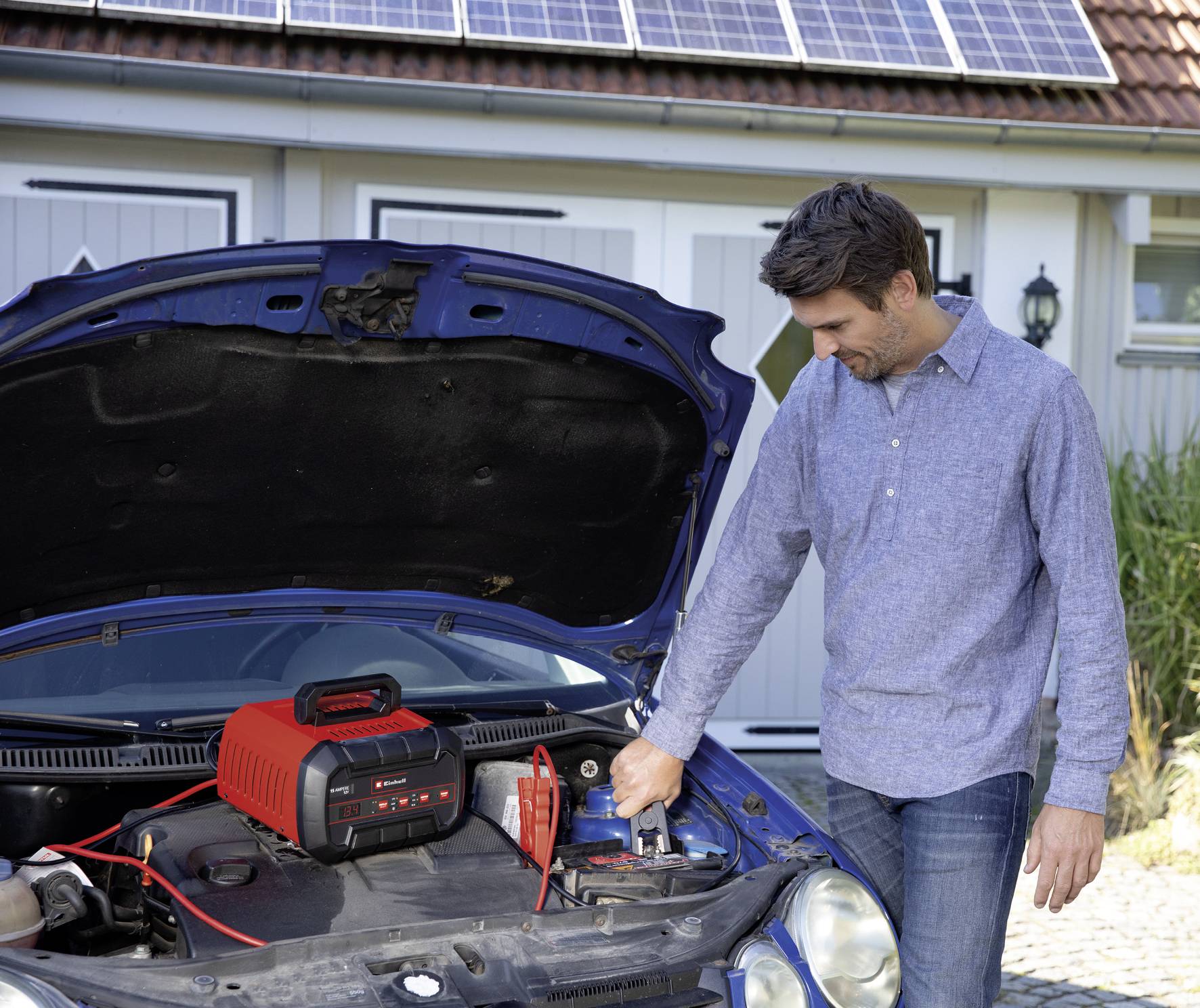 Muž nabíjí autobaterii pomocí nabíječky. Auto stojí s otevřenou kapotou před garáží s instalovanými solárními panely.