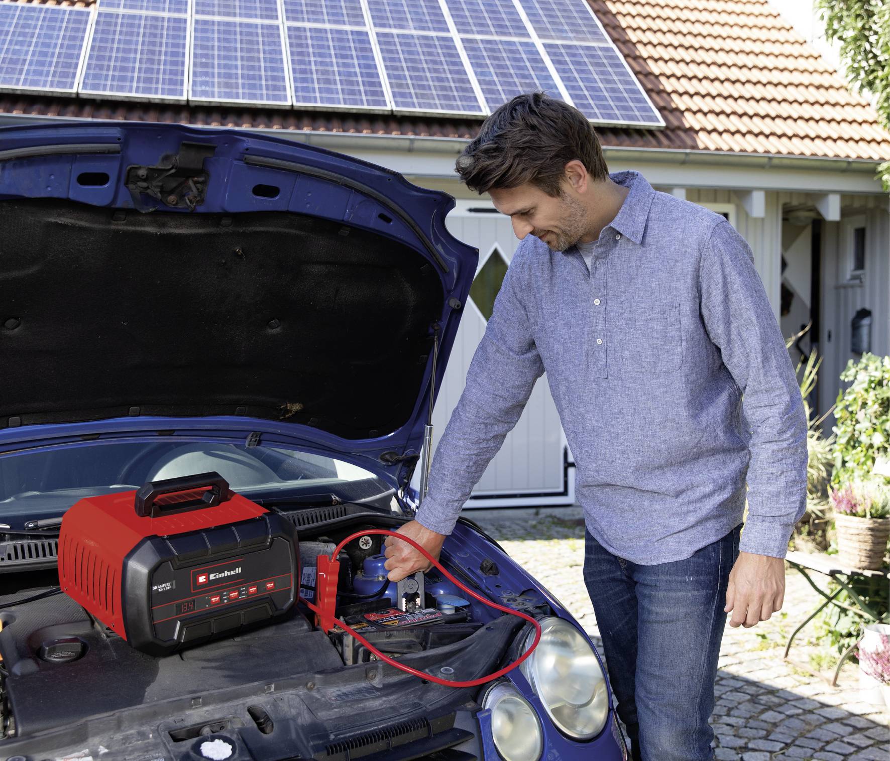 Muž nabíjí automobil startovacím zařízením, přičemž v pozadí je vidět dům se solárními panely na střeše.