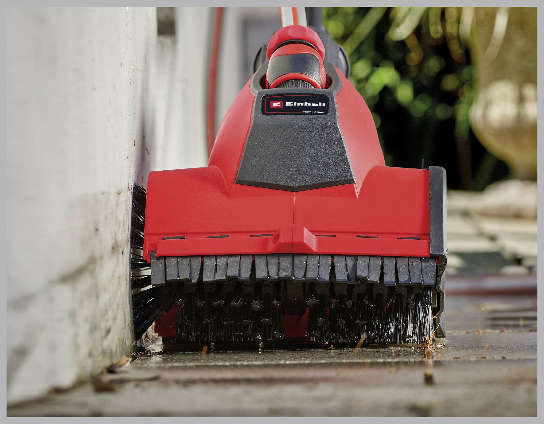 Červený elektrický kartáčový vysavač čistí terasu podél betonové zdi, odstraňuje špínu a plevel mezi spárami.