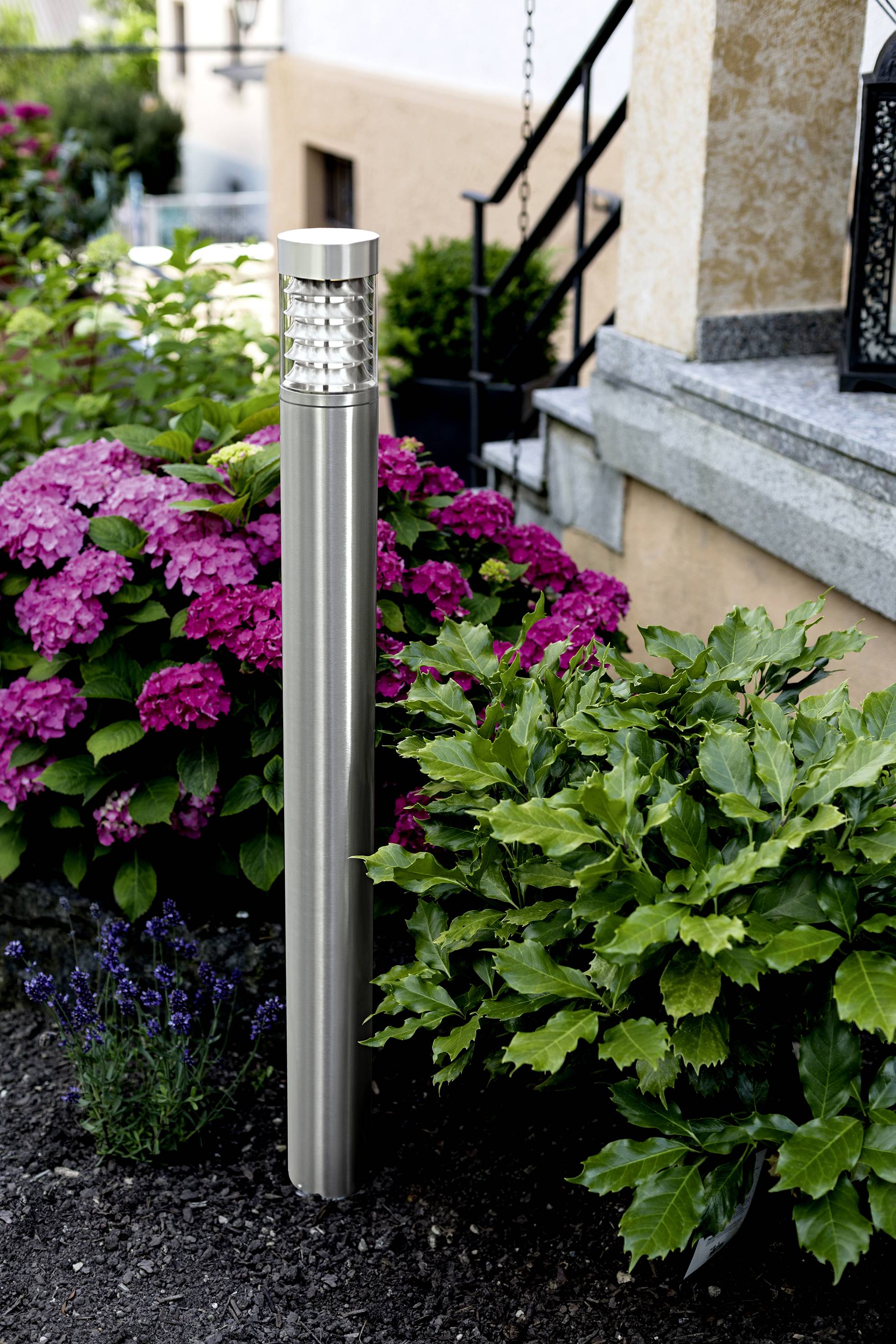 Nerezová lampa v zahradě osvětluje barevné hortenzie a zelené keře před kamenným schodištěm.
