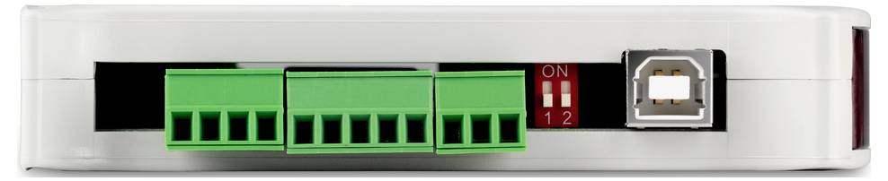 Elektronické zařízení s USB portem, dvěma zelenými připojovacími svorkami a červenými přepínači. Zařízení stojí na bílém povrchu.