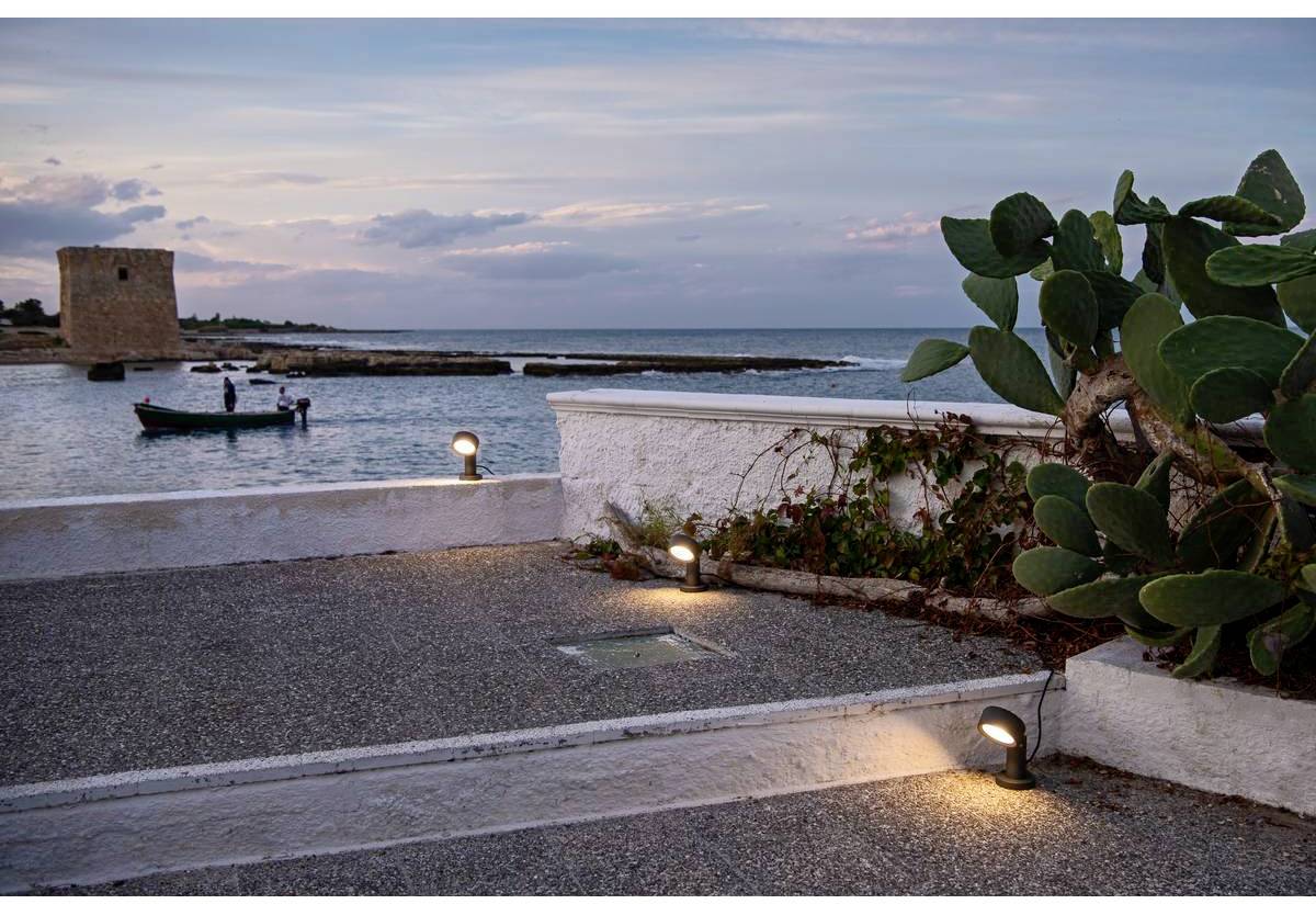 Pobřeží při západu slunce se starým věžovým majákem, loď na vodě a kaktusy v popředí. Tři osoby v lodi, klidná atmosféra.