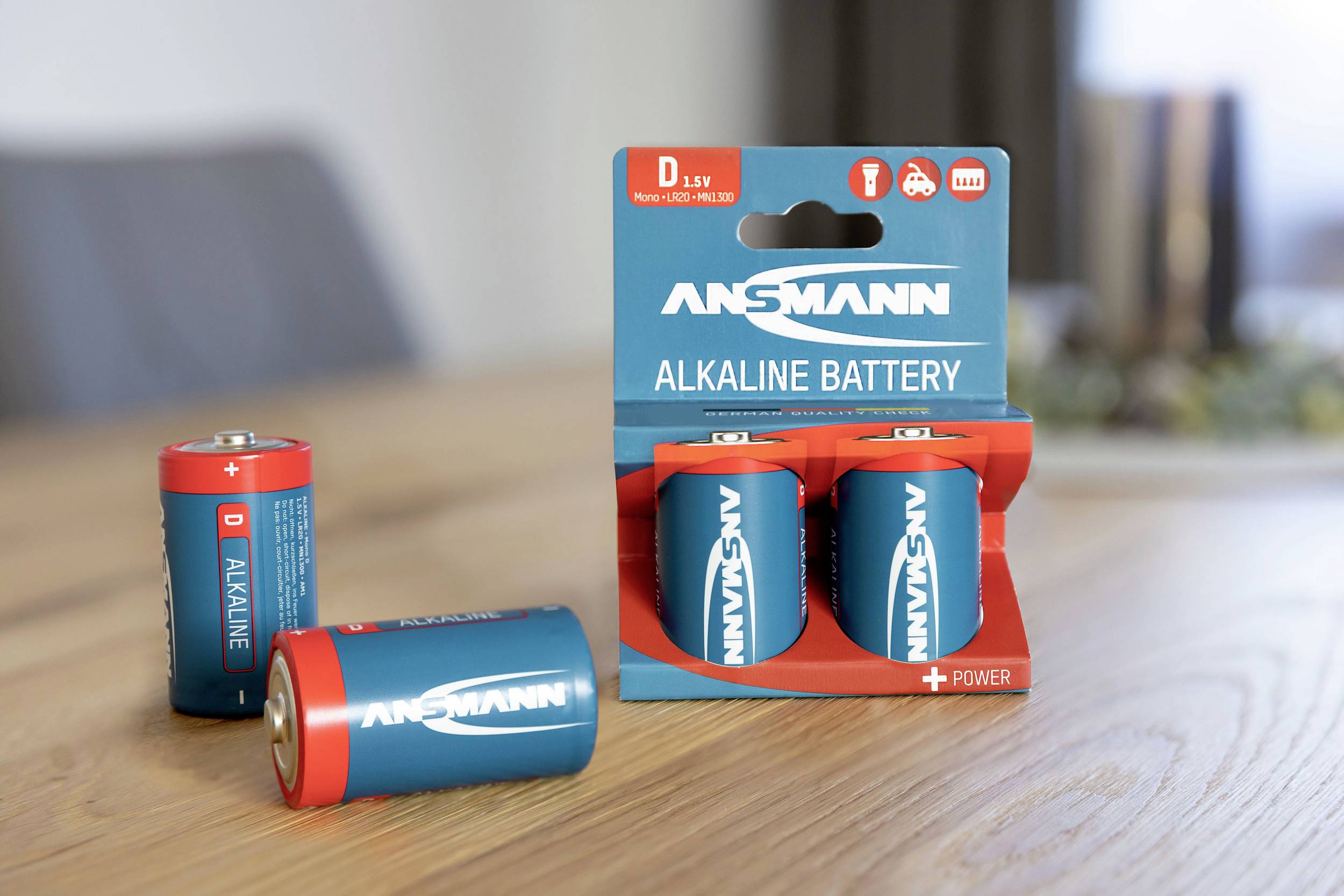 Čtyři modré alkalické baterie Ansmann, typ D, na stole; dvě stojící v originálním obalu, dvě volně ležící.