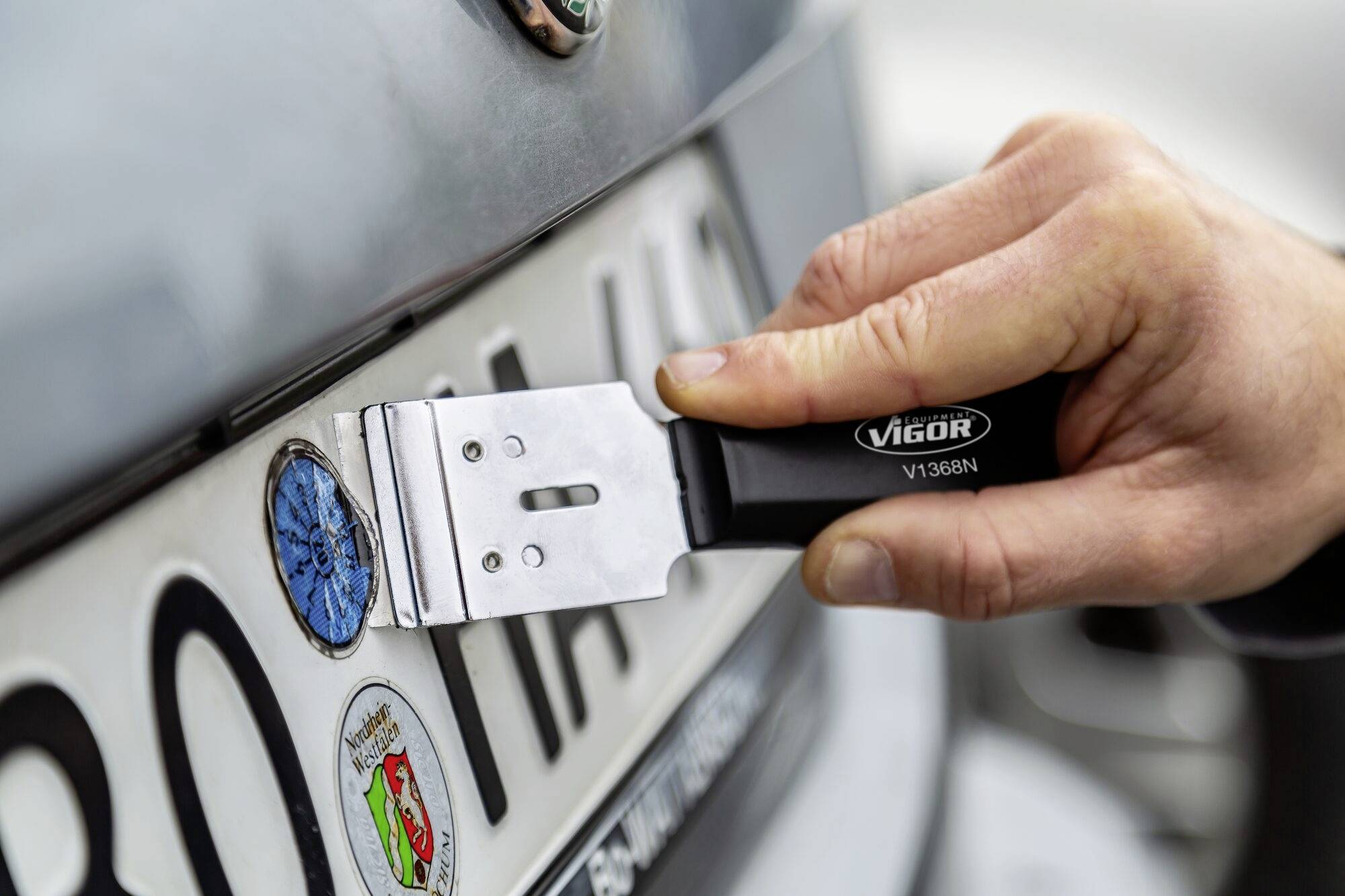 Ruka škrábe škrabkou Vigor modrý TÜV štítek z šedé registrační značky; záběr je zaměřen na samotnou akci.