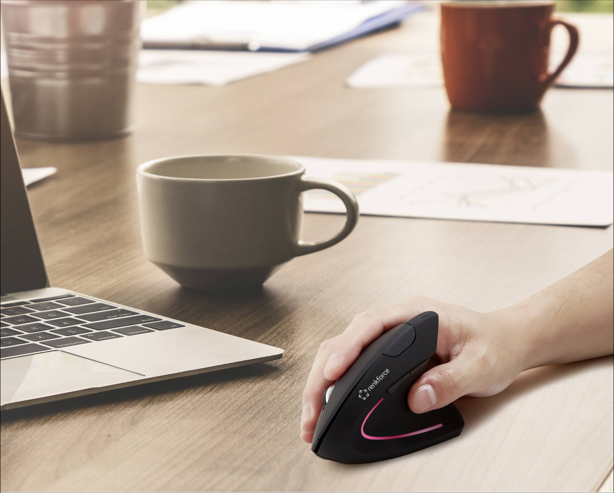 Ruka drží ergonomickou vertikální myš u laptopu na stole s kávovými šálky v pozadí, což symbolizuje pohodlnou práci.