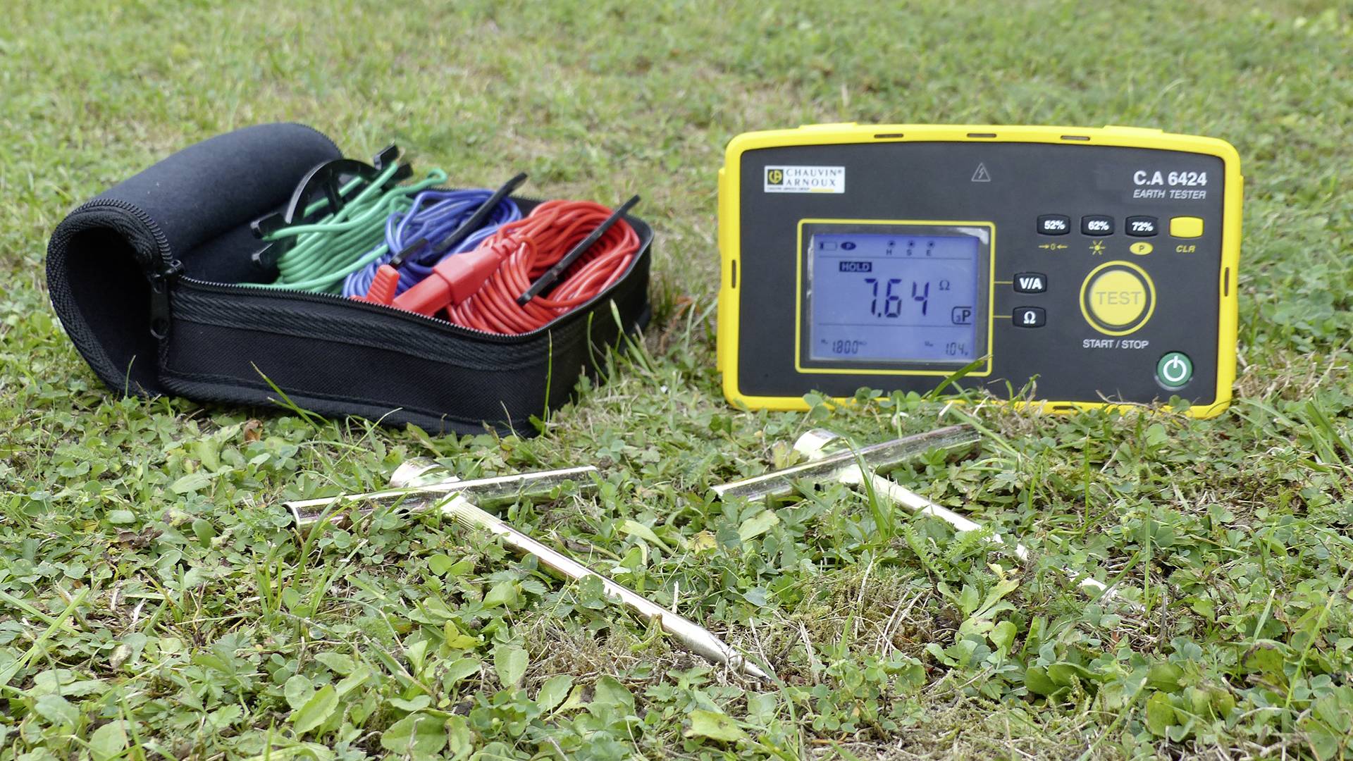 Měřicí přístroj pro měření zemního odporu s displejem zobrazuje '7,64 Ω'. Vedle přístroje leží měřicí kabely a sondy na travnaté ploše.