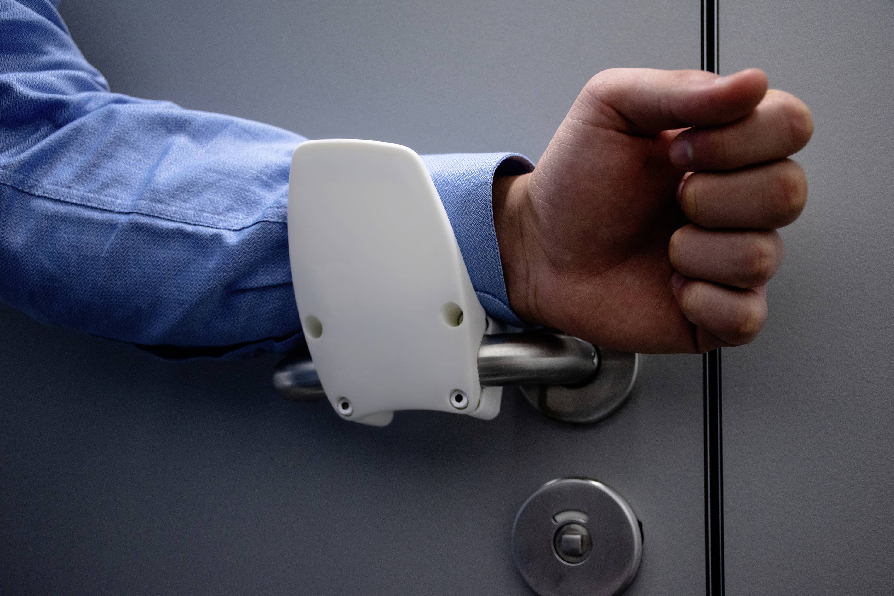 Detailní záběr paže v modrém tričku, která otevírá dveře. Bílá plastová klička je nasazena kolem zápěstí, aby se zvýšila hygienická bezpečnost.