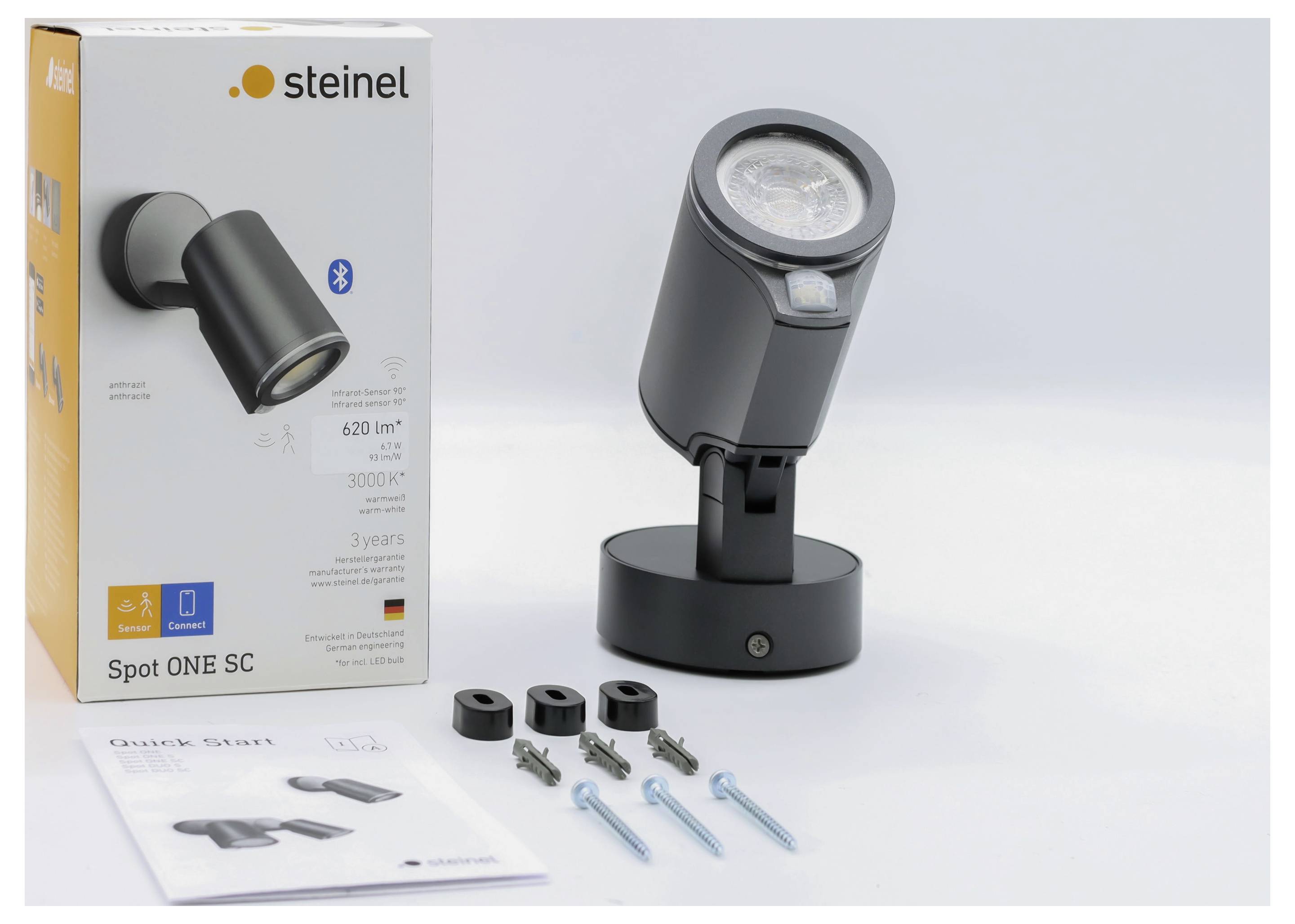 Produktová fotografie chytré reflektorové lampy Steinel Spot ONE SC s Bluetooth, jejíž balení, průvodce rychlým startem a montážní hardware jsou zobrazeny na bílém pozadí.
