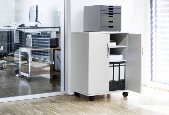 Kancelář s otevřenou skříní plnou složek v popředí a stolem s počítačem v pozadí.