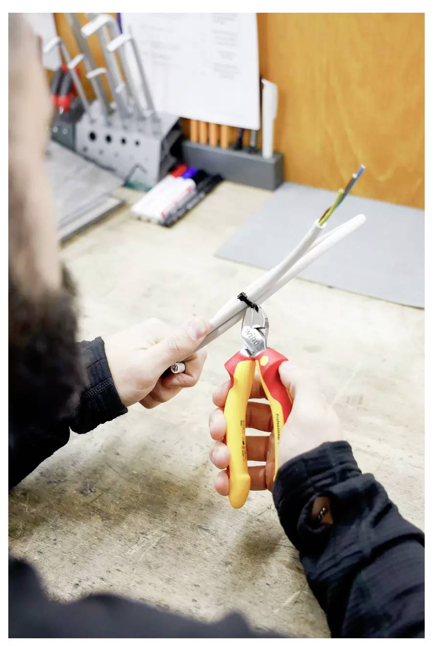 Osoba používá nůžky na drát k obnažení kabelu na pracovním stole. V pozadí se nacházejí různé nástroje a tužky.