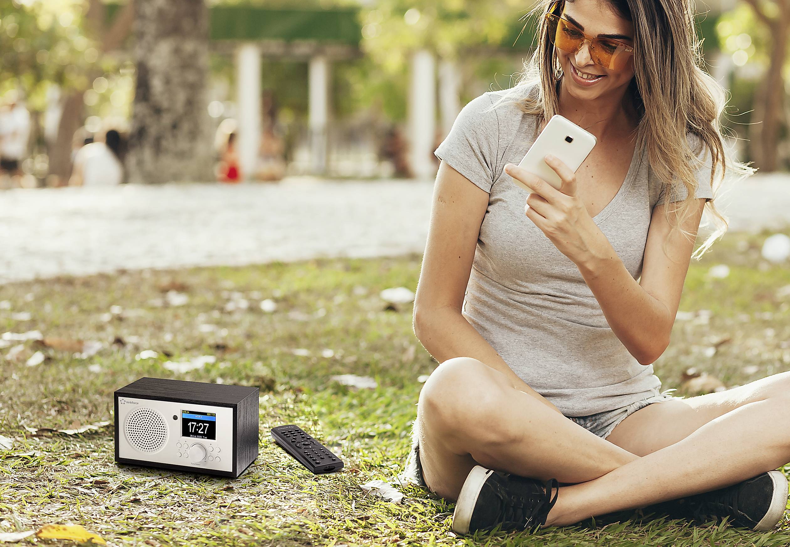 Žena sedí venku na trávě a dívá se na svůj smartphone. Vedle ní stojí rádio s digitálním displejem.