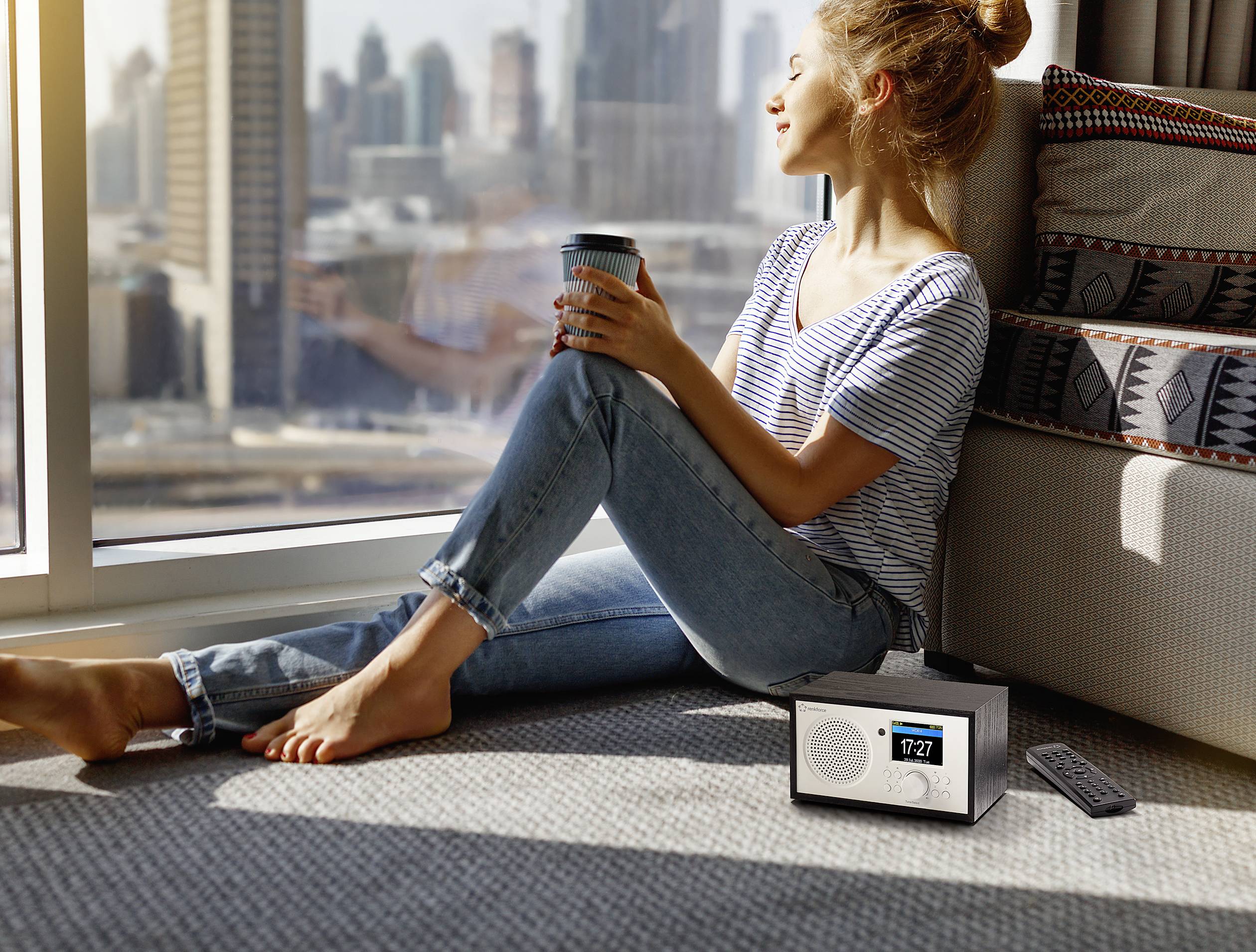 Žena sedí u okna s úsměvem, drží šálek a užívá si výhled na město. Vedle ní stojí rádio s viditelným časem.