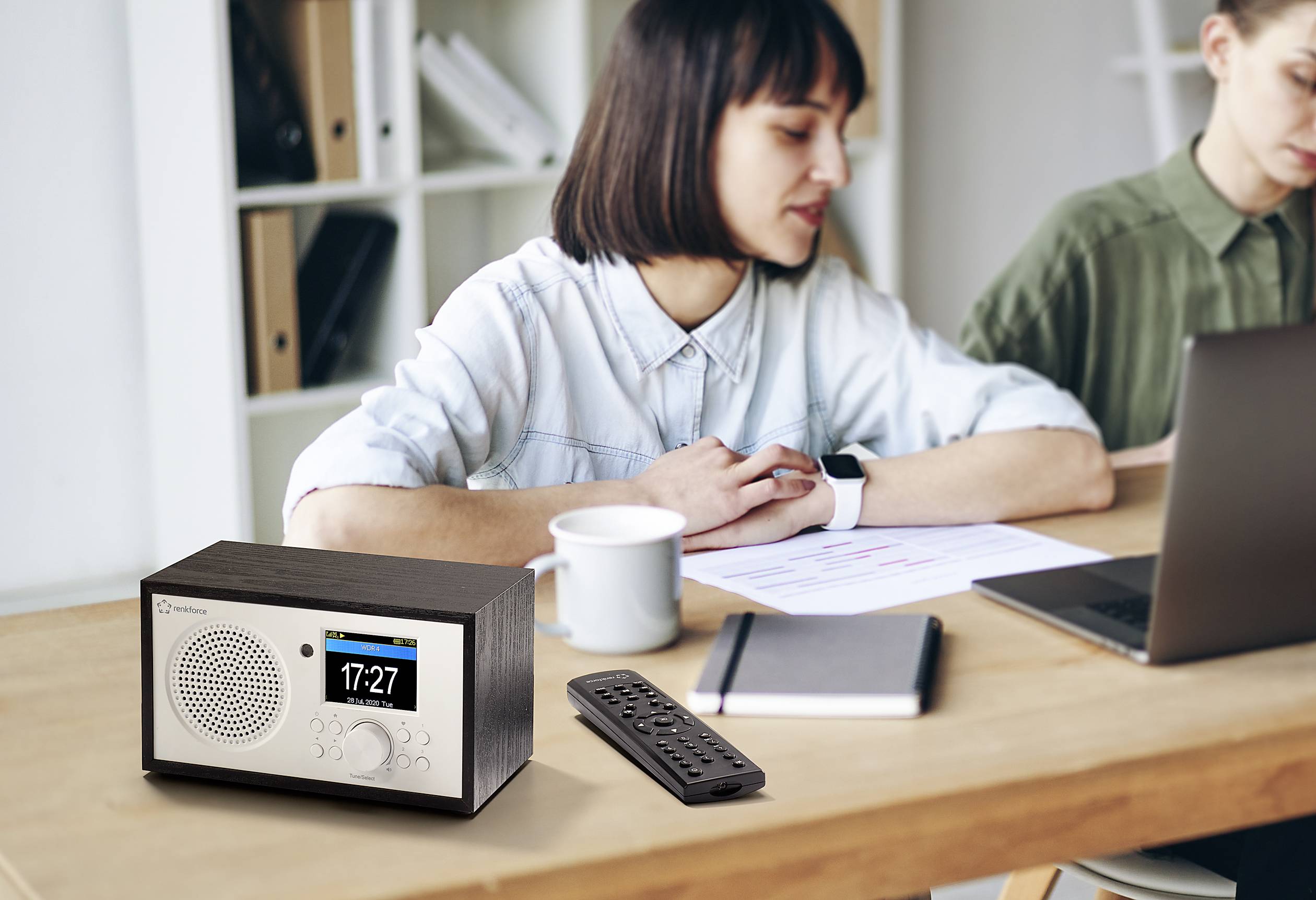 Žena sedící u stolu pracuje na notebooku vedle druhé ženy. Na stole leží rádio a dálkový ovladač.