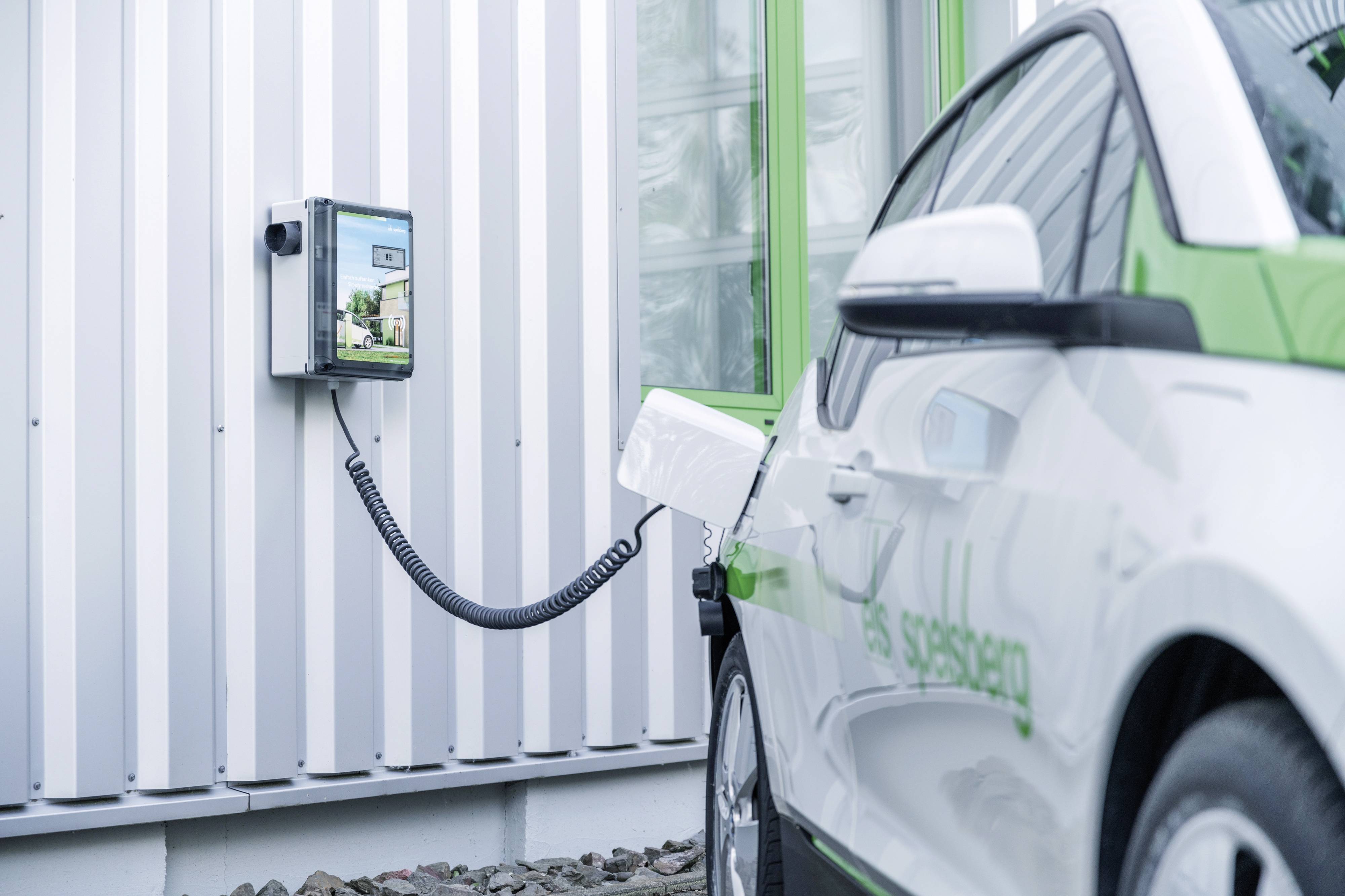 Bílý elektromobil se nabíjí u nabíjecí stanice namontované na stěně moderní budovy.