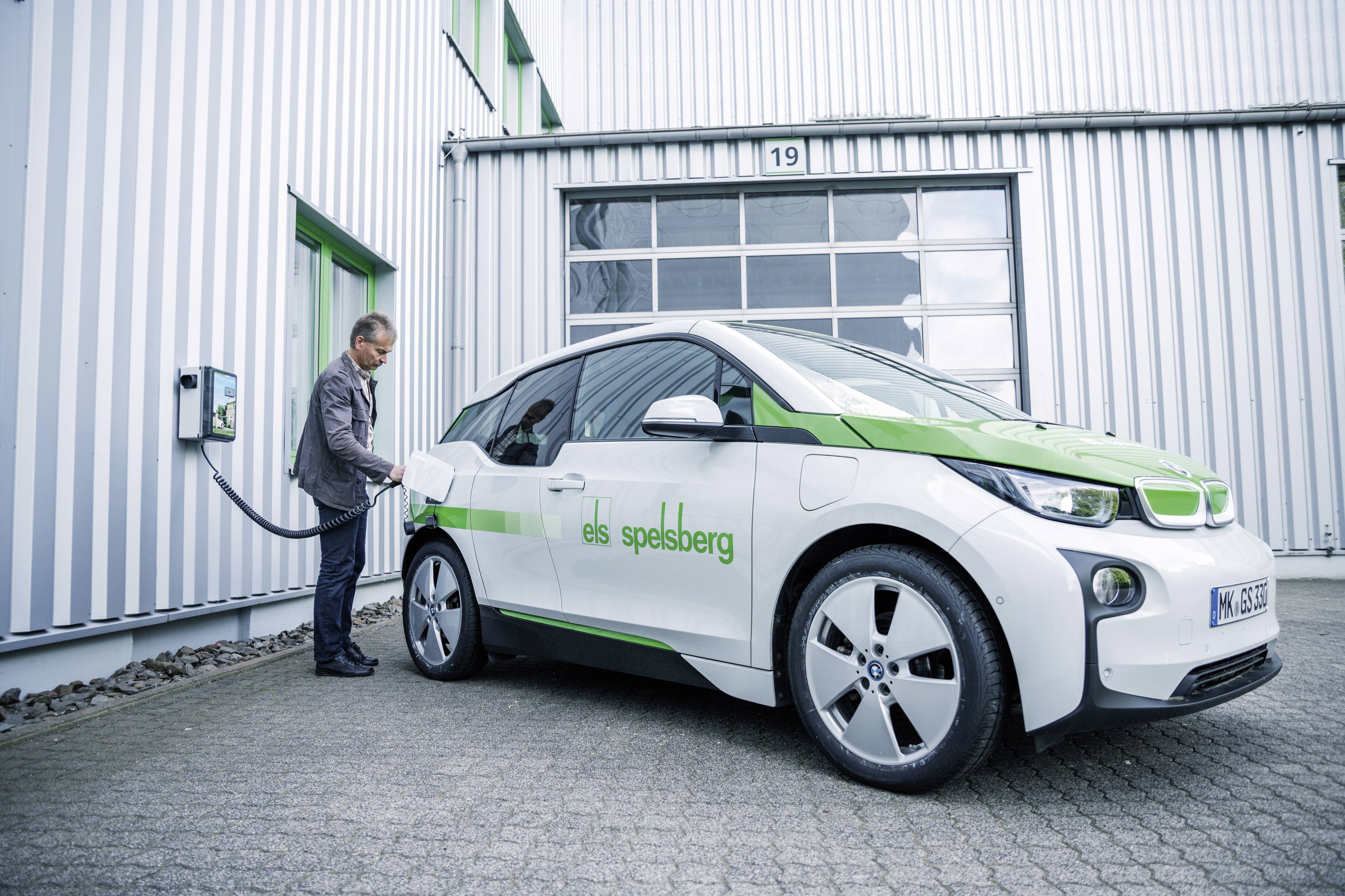 Muž nabíjí bílé elektrické auto s zeleným pruhem na nabíjecí stanici před průmyslovým objektem.
