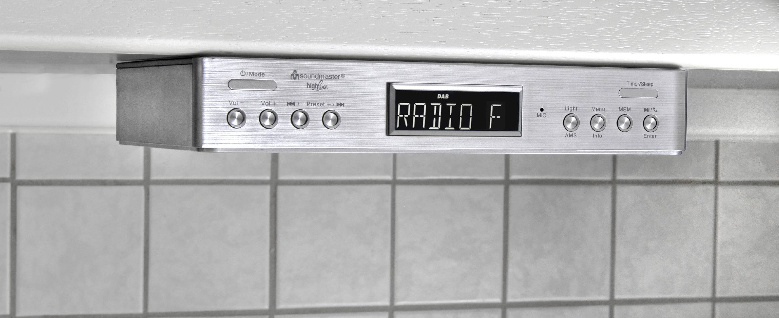 Rádio namontované pod kuchyňskou policí zobrazuje 'RADIO F' na displeji, s tlačítky pro výběr stanice, nastavení hlasitosti a dalších funkcí. V pozadí šedé obkladačky.
