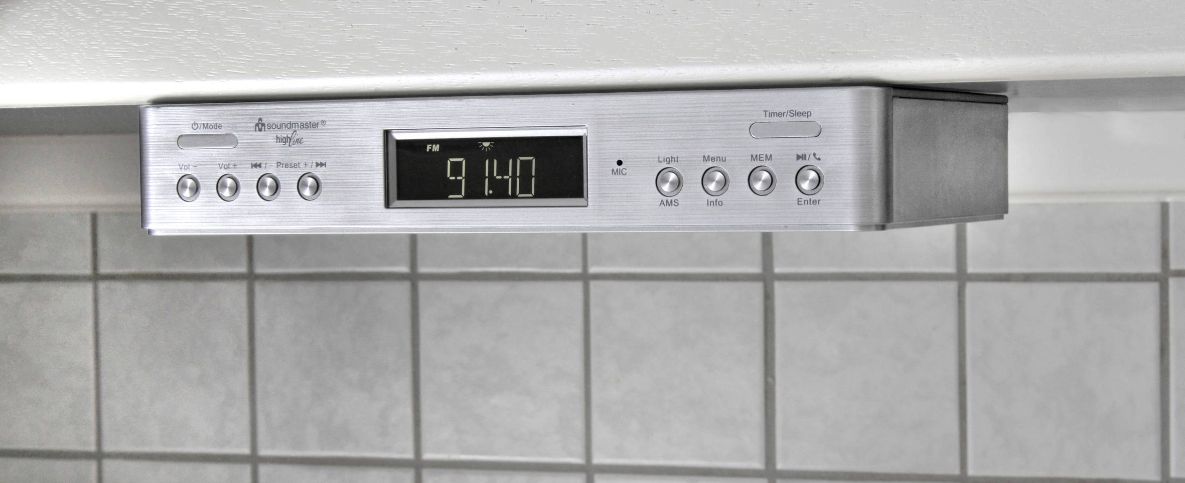 Elektronické hodiny ve skříni z nerezové oceli ukazují 9:14. Obklopeny ovládacími tlačítky pro světlo a digestoř nad obloženou stěnou.