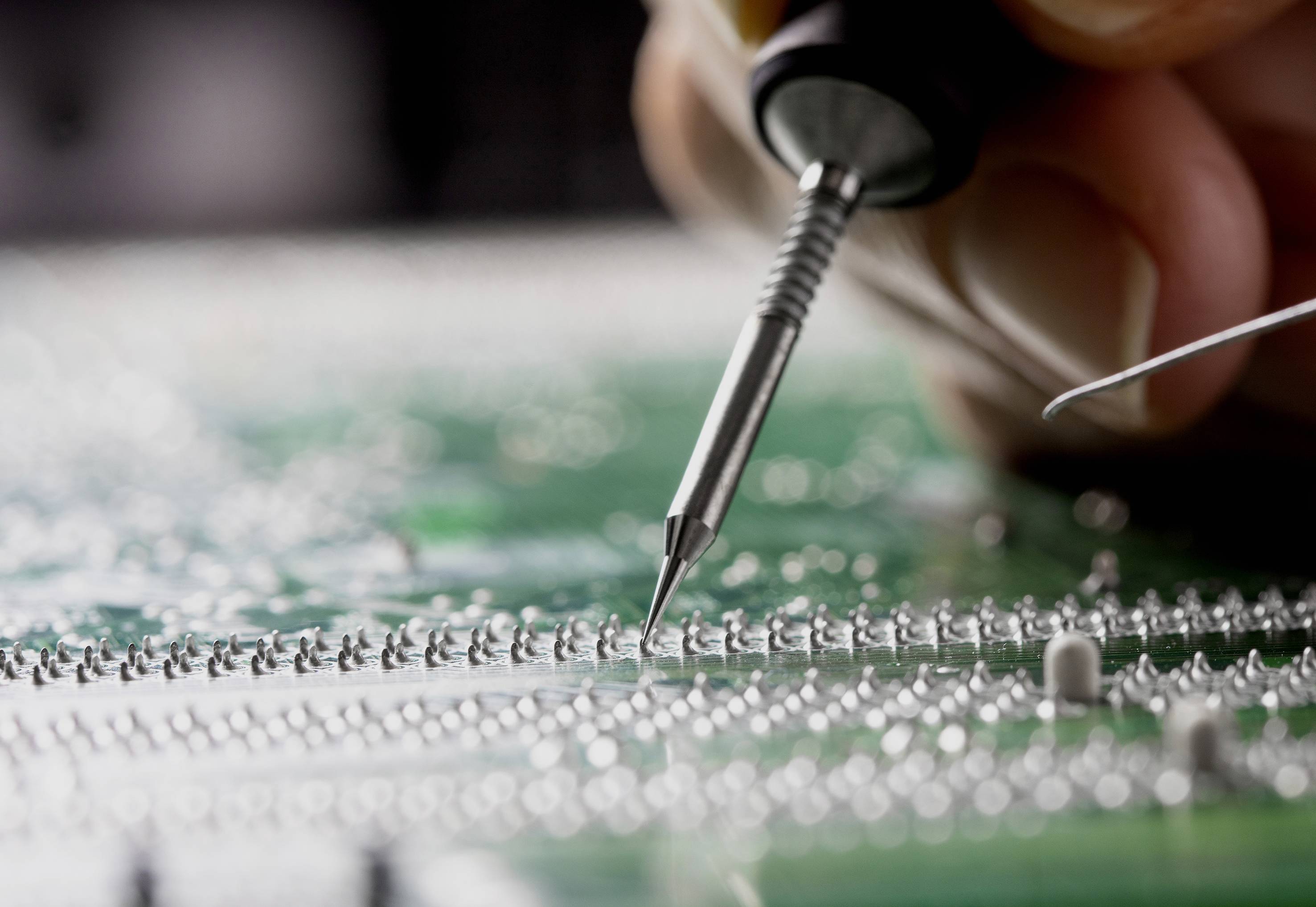 Detailní záběr ruky, která pájecím přístrojem pracuje na zelené desce plošných spojů. Důraz je kladen na přesnost a technologii.