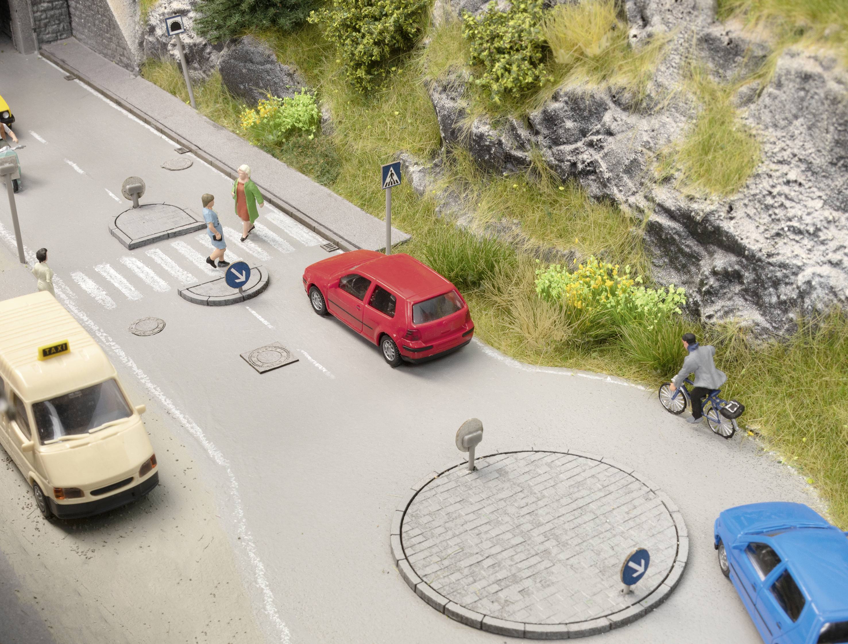 Model silnice s miniaturními auty, přechodem pro chodce, červeným autem a cyklistou jedoucím po cyklostezce podél skalnatého svahu.