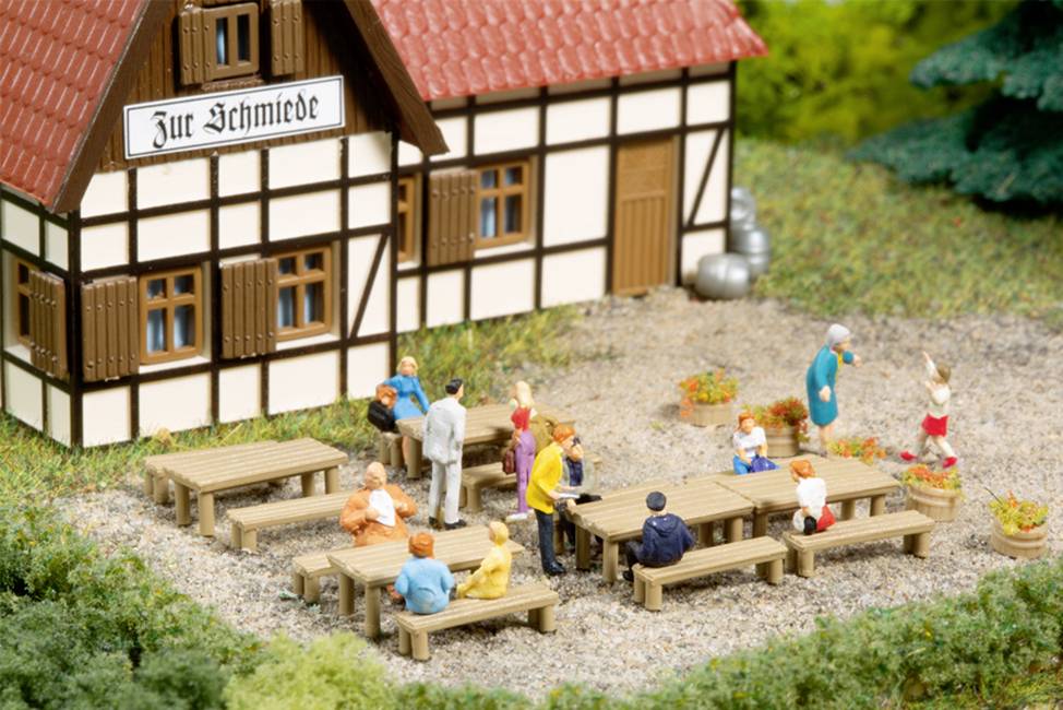 Miniaturní model zobrazuje německý hrázděný dům s cedulkou 'U kovárny'. V popředí sedí figurky na dřevěných lavicích a stolech venku.
