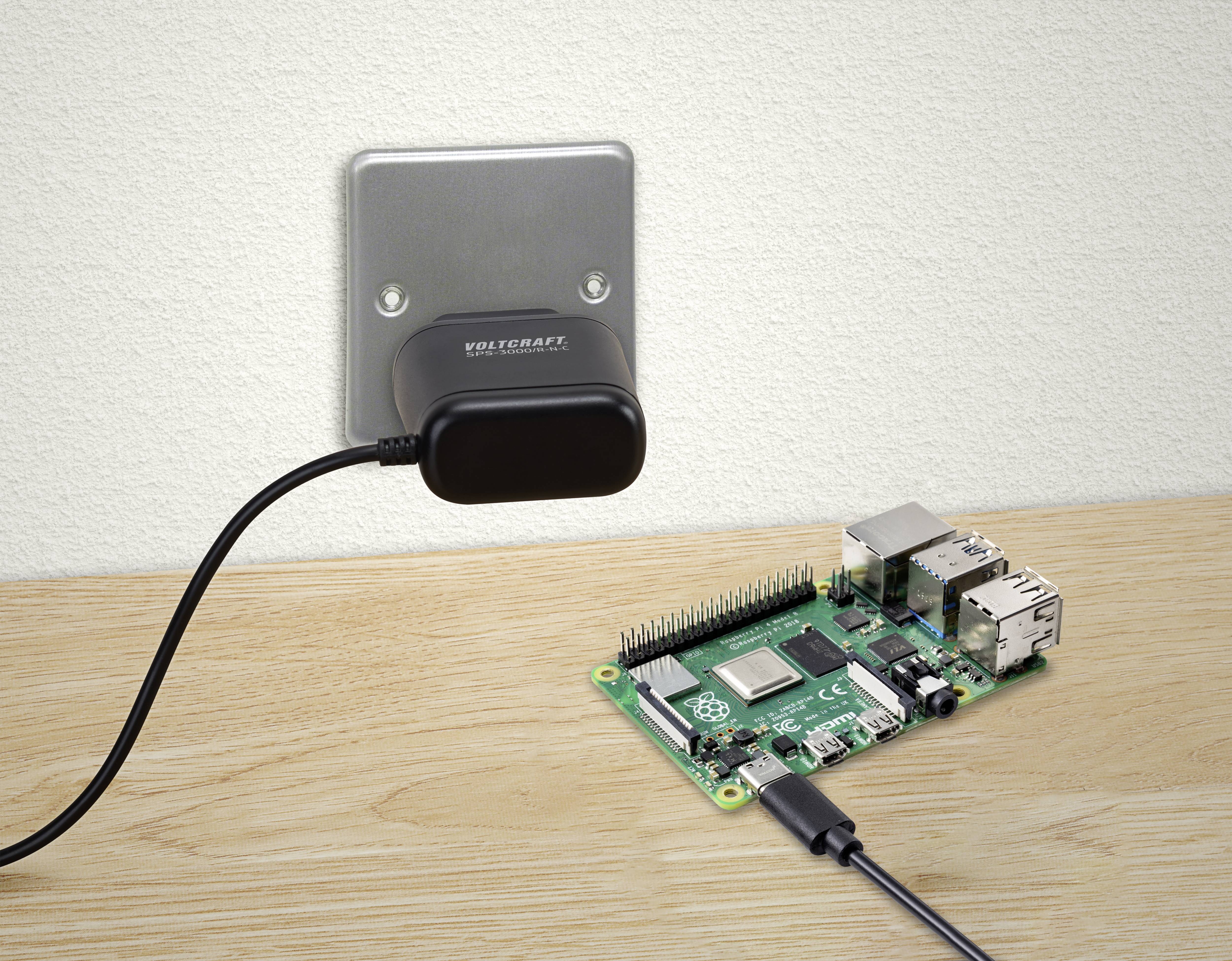 Na dřevěném stole je vidět Raspberry Pi, připojené k napájecímu adaptéru zapojenému do elektrické zásuvky.
