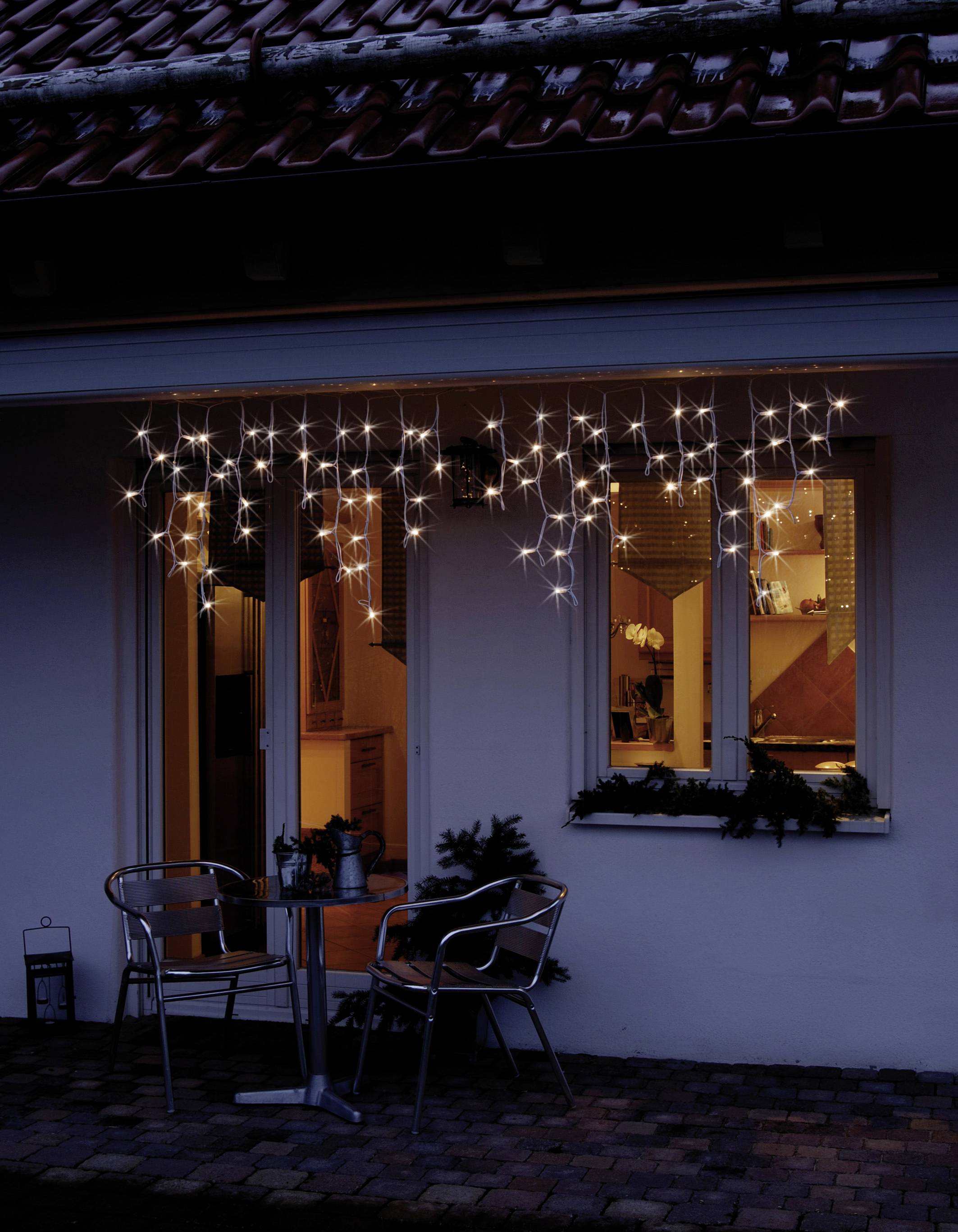 Osvětlený vchod do domu s vánoční výzdobou. Světelný řetěz v okně a malé posezení před ním. Útulná večerní atmosféra.