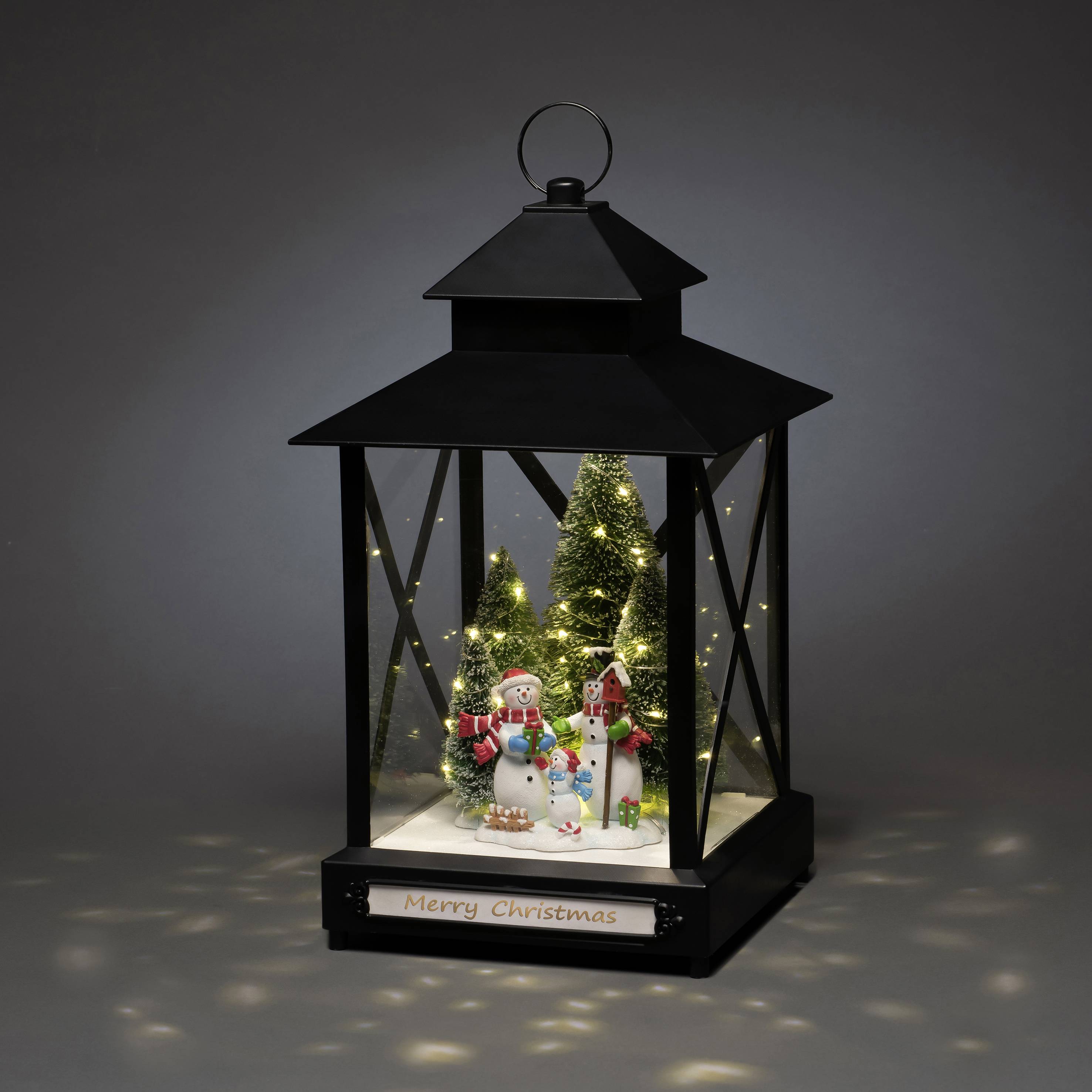 Černá lucerna s dekorací sněhuláka a vánočními stromky uvnitř pod LED osvětlením. Nápis 'Merry Christmas'.