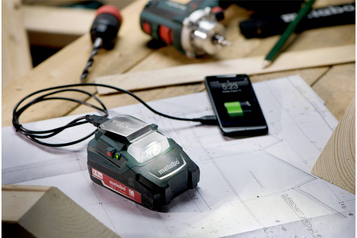 Nabíjecí stanice pro akumulátory elektrického nářadí na dřevěném stole, propojená se smartphonem, zobrazuje průběh nabíjení. V pozadí leží stavební plány a nástroje.