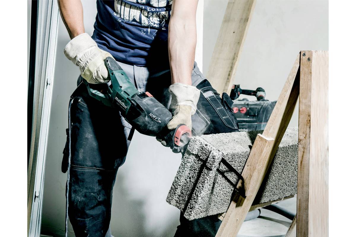 Řemeslník řeže elektrickou pilou beton. Má na sobě pracovní rukavice a ochranné kalhoty. Je patrná atmosféra staveniště.