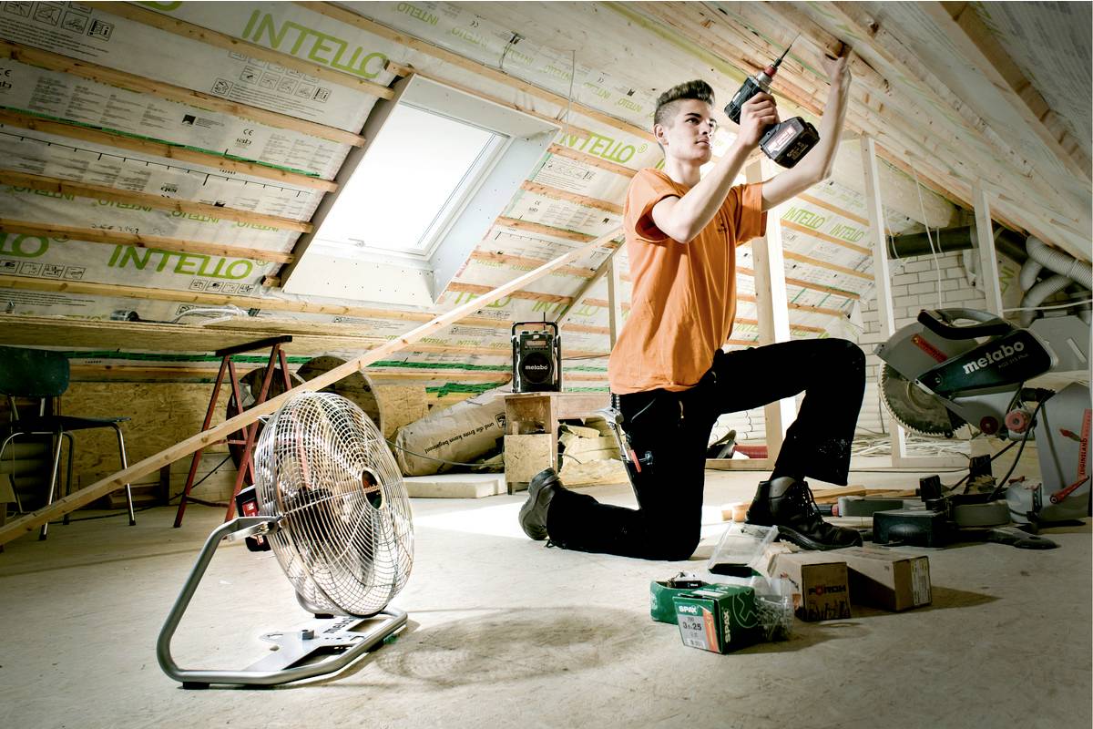 Řemeslník klečí na půdě a pracuje s elektrickým nástrojem. V popředí stojí ventilátor.