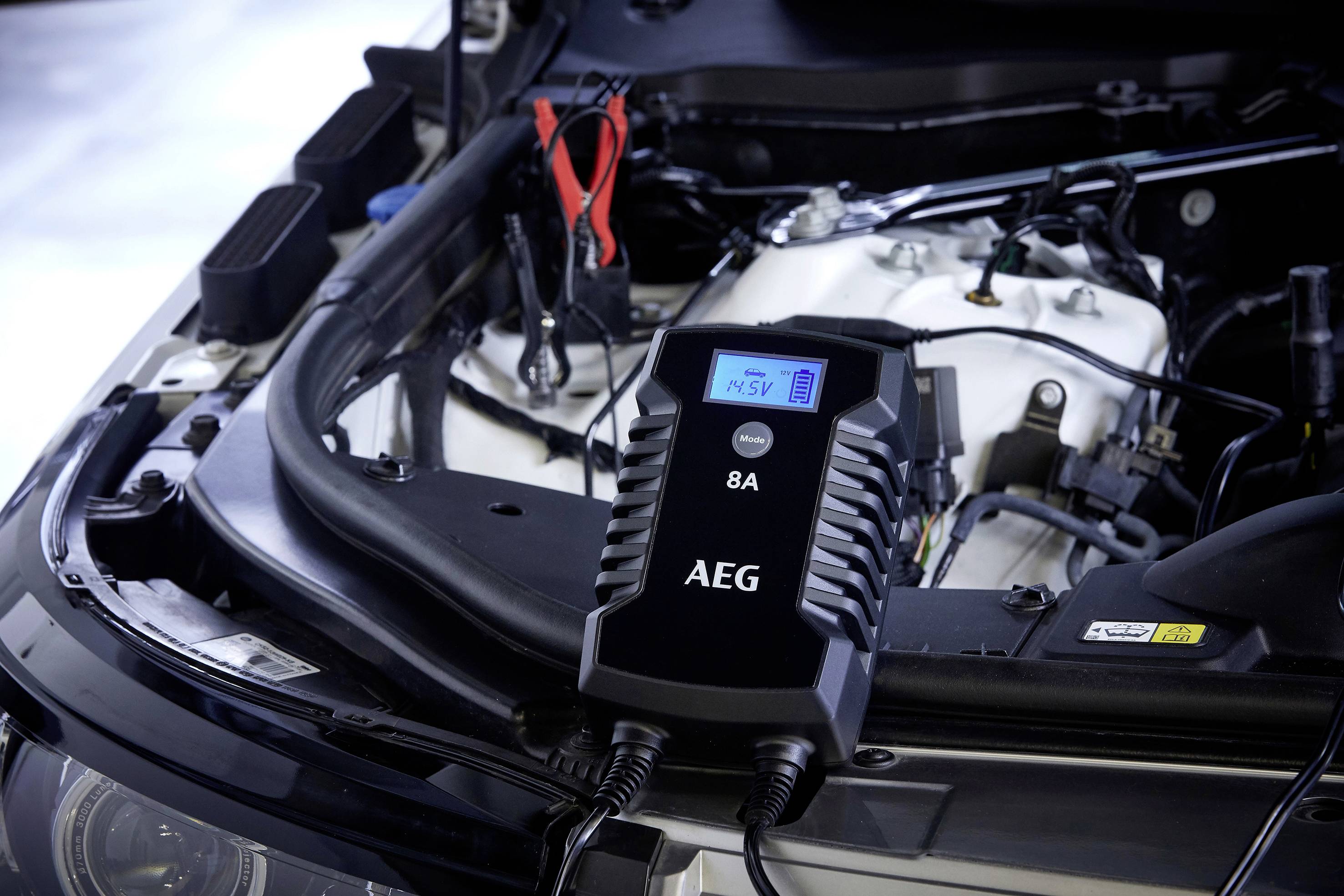 Nabíječka baterií značky AEG, připojená pod otevřenou kapotou automobilu. Stav nabití je zobrazen na displeji.