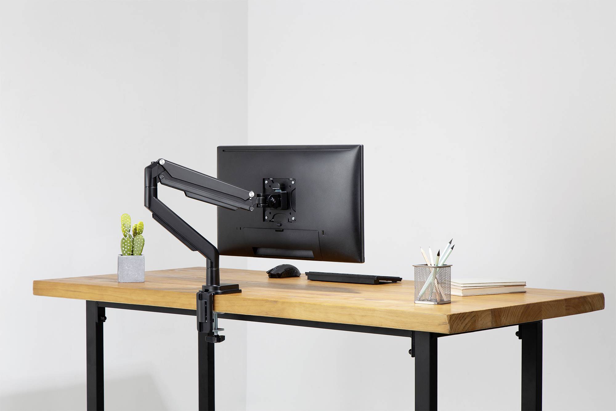 Moderní psací stůl s dřevěnou deskou a černou kovovou konstrukcí. Na stole stojí monitor s kloubovým ramenem, malý kaktus a tužky.