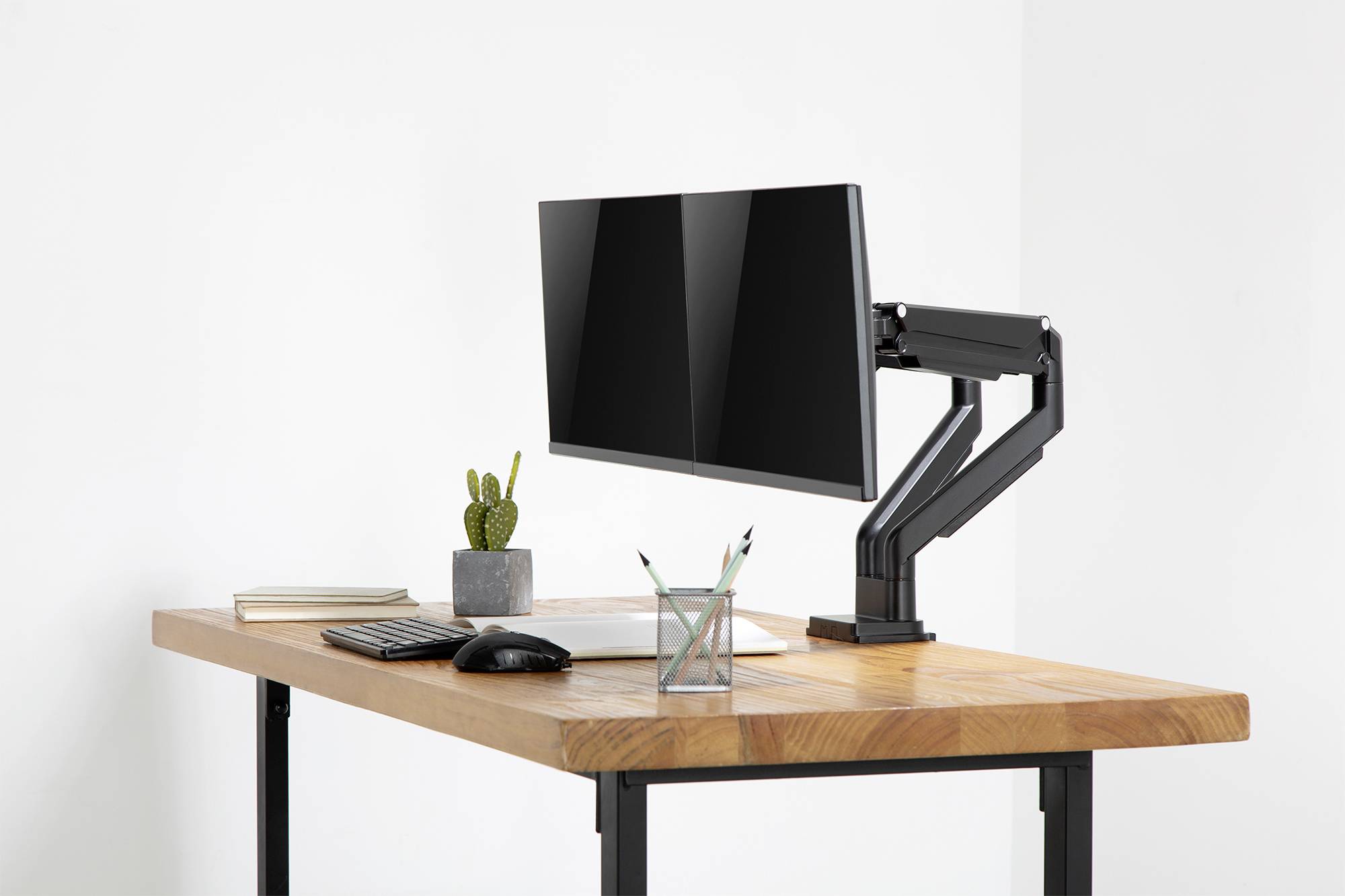 Moderní psací stůl s monitorem na nastavitelném rameni, vedle kterého jsou klávesnice, myš, poznámkový blok, stojánek na tužky a kaktus.